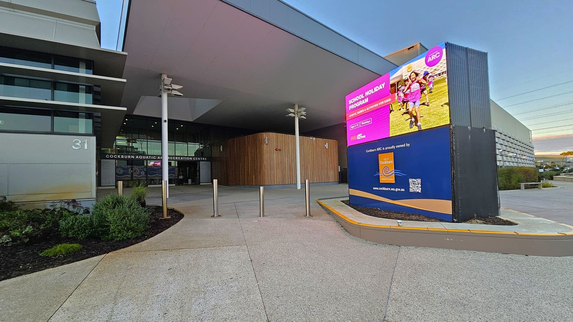 Entrance to a modern building with a large digital sign on the right displaying a school holiday programme advertisement. The building has a sleek design with glass and metal elements. The entrance is marked by the words "Cockburn Aquatic and Recreation Centre" above the doors. There are several bollards and a landscaped area with shrubs on the left. The ground is paved with light-coloured concrete, and the ceiling features a large overhang.
