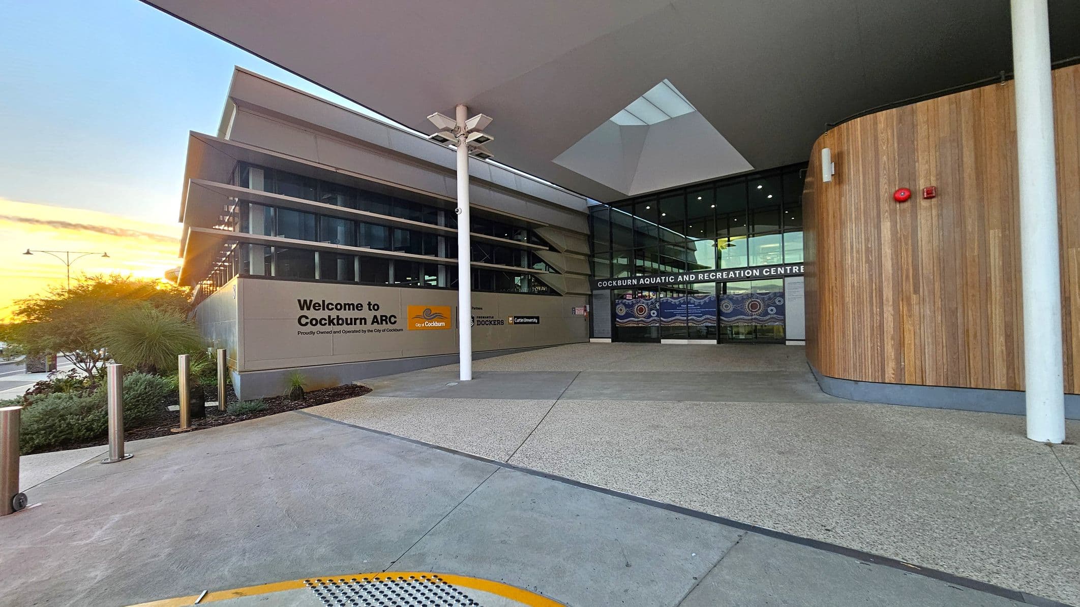 Entrance to a modern building with a sign reading "Welcome to Cockburn ARC" on the left. The structure features large glass windows and a wooden panelled wall on the right. A white pillar stands in the centre, supporting a canopy with geometric shapes. The ground is paved with textured concrete, and there are landscaped plants near the entrance. The building's name, "Cockburn Aquatic and Recreation Centre," is displayed above the glass doors.