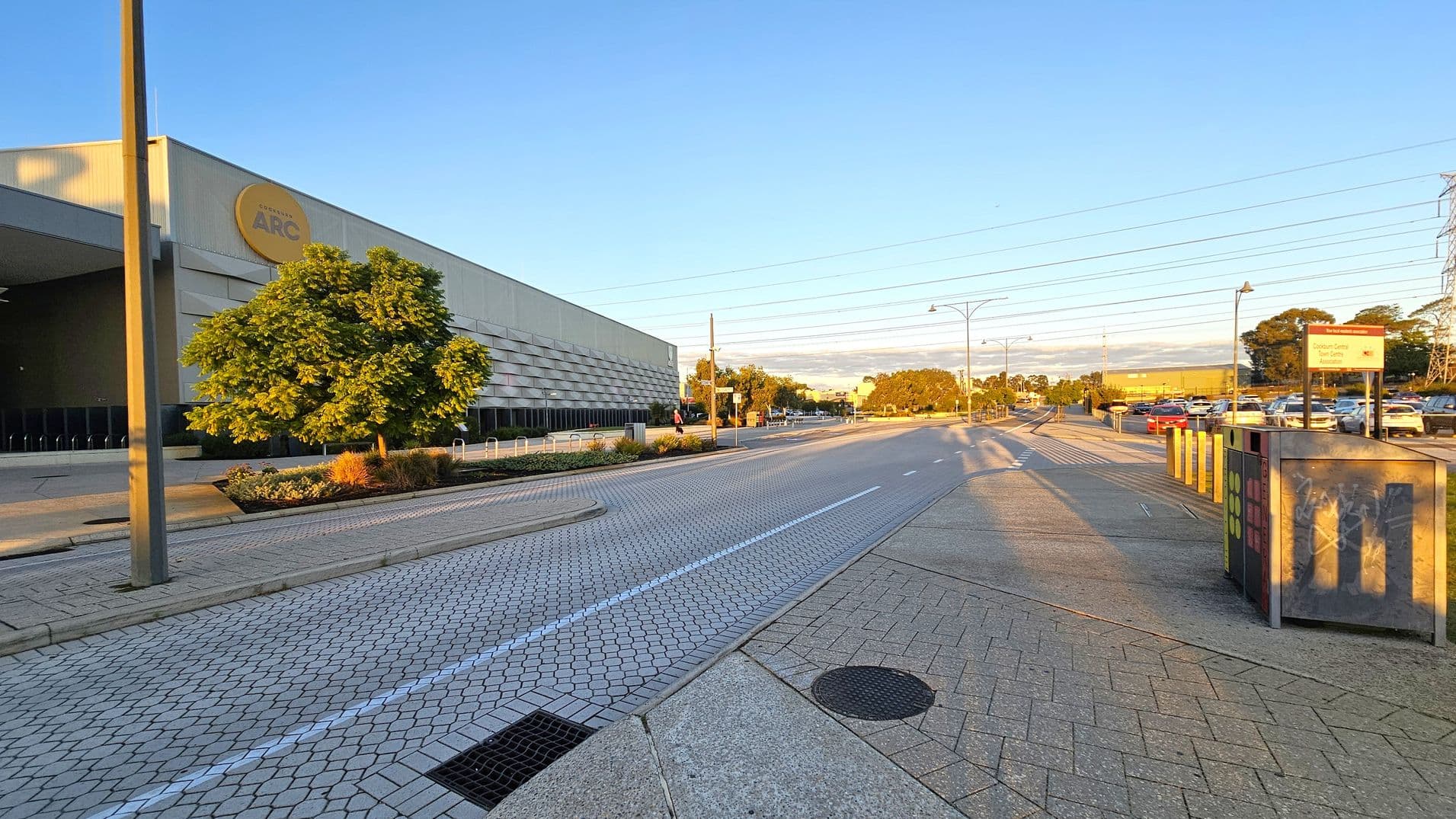 A wide street with a hexagonal-patterned pavement runs through the centre. On the left, a large building with a sign and a tree in front. On the right, a car park with several vehicles and a sign. A utility box with graffiti is near the foreground. The road is lined with streetlights and power lines. The pavement is a mix of concrete and patterned tiles. Trees and shrubs are visible in the background.