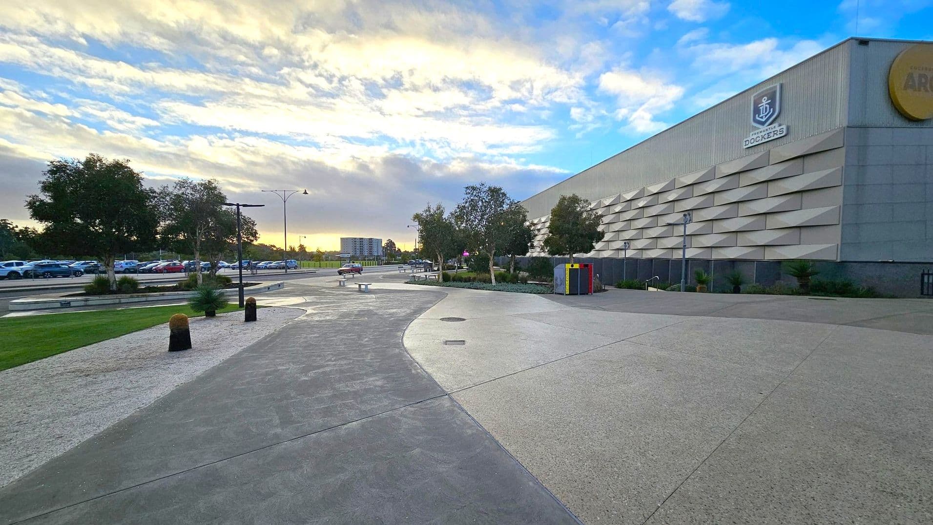 A wide pathway curves through the foreground, bordered by a grassy area with trees on the left. Several parked cars are visible in the distance. On the right, a large building with a textured facade and a logo is present. A few benches and a colourful bin are near the building. The background features more trees and a distant building under a partly cloudy sky.