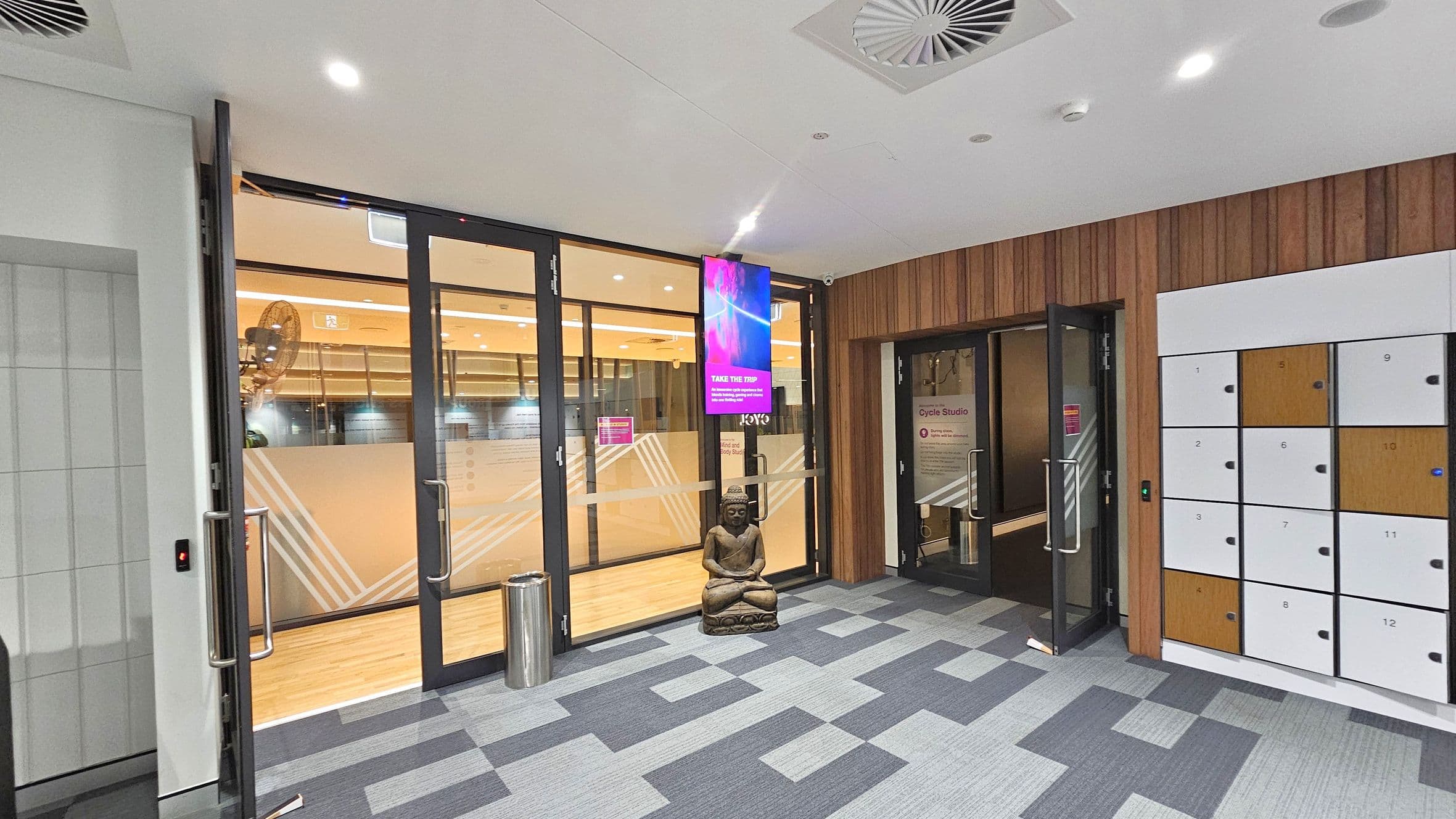 A modern interior space with a patterned carpet floor. On the left, there are glass doors with metal handles leading to another room, featuring frosted glass with geometric designs. A digital screen is mounted above the doors. In the centre, a statue of a seated figure is placed on the floor. On the right, there is a wall of lockers with numbered doors in white and wood tones. The ceiling is white with recessed lighting and air vents.