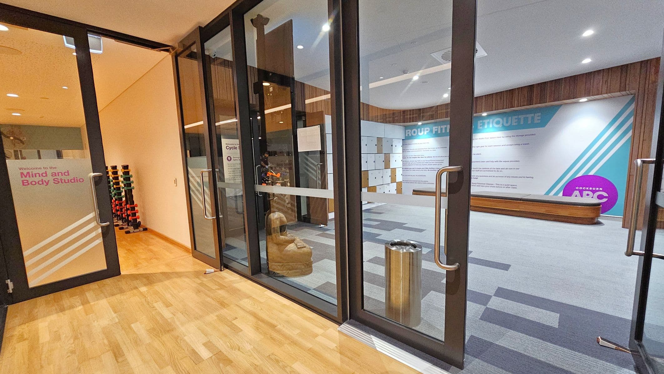 On the left, there is a glass door with "Welcome to the Mind and Body Studio" written on it, leading to a room with a wooden floor and a rack of colourful dumbbells. On the right, another glass door opens into a space with a carpeted floor, featuring a wall with "GROUP FITNESS ETIQUETTE" text and a bench. A metal bin is near the door. The ceiling is white with recessed lighting, and the walls have a mix of wood panelling and painted surfaces.