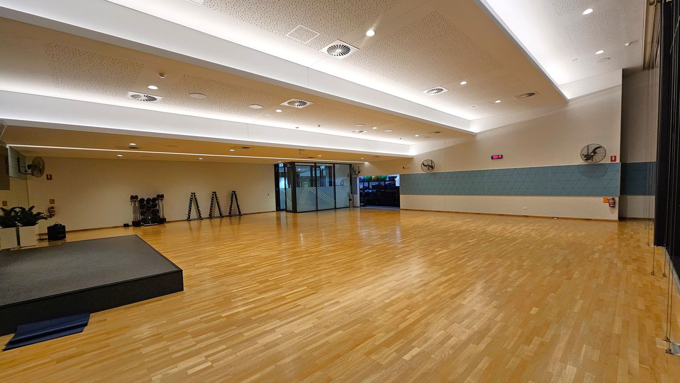 A spacious indoor room with a wooden floor and a high ceiling featuring recessed lighting and air vents. On the left, there's a small stage with a dark surface and a stack of exercise mats. In the centre, a glass door leads to another area. On the right, the wall has a blue panel with mounted fans and a digital clock displaying "5:31". The room is equipped with exercise equipment like dumbbells and step platforms against the far wall.