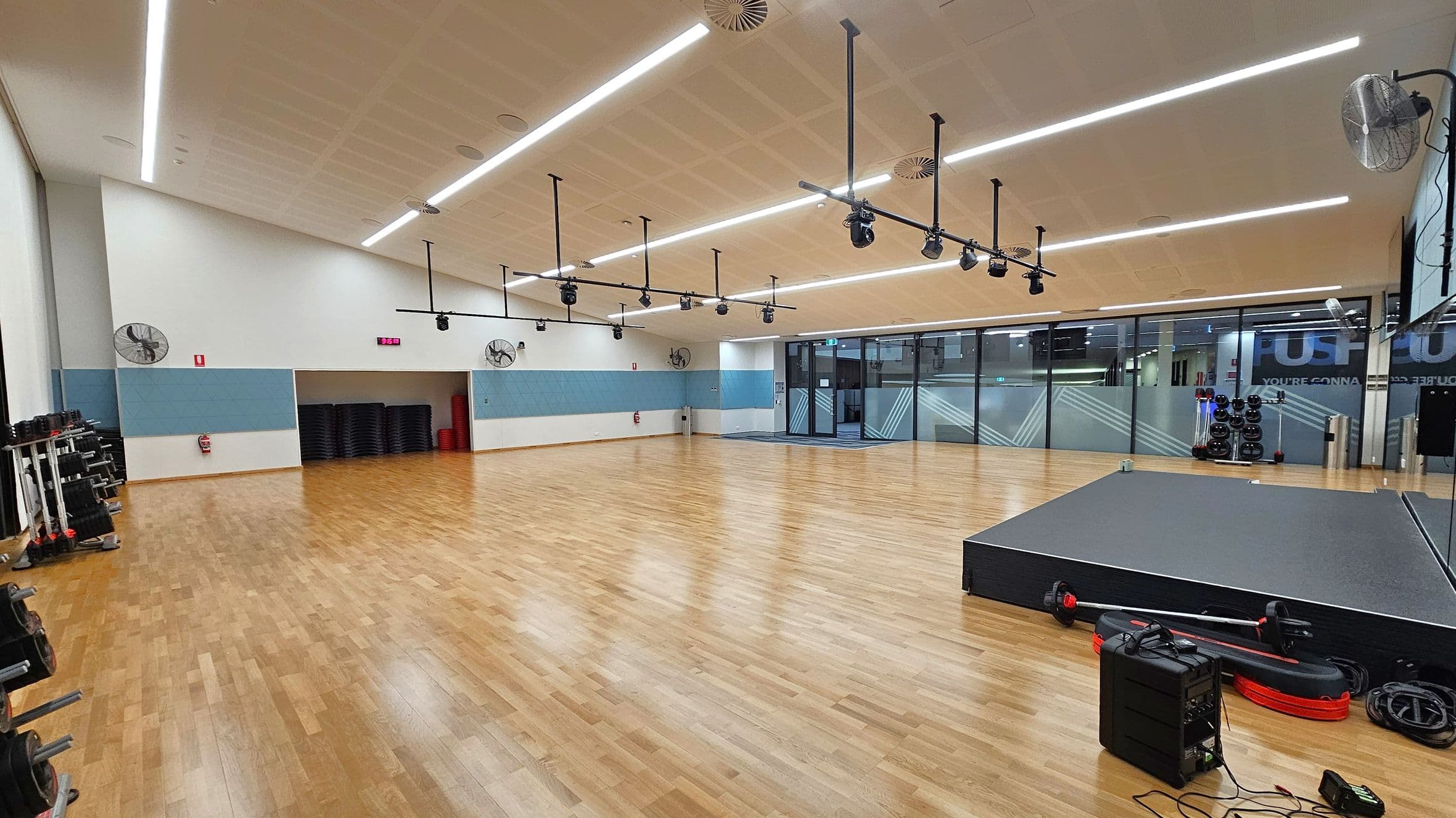 A spacious gym studio with a wooden floor and a high ceiling featuring linear lights. On the left, there are weight racks and a stack of black mats against a light blue and white wall. The centre has a digital clock and wall-mounted fans. On the right, a raised platform with exercise equipment and cables is visible. The back wall is made of glass panels with frosted designs, and there are more weights and exercise balls near the glass.
