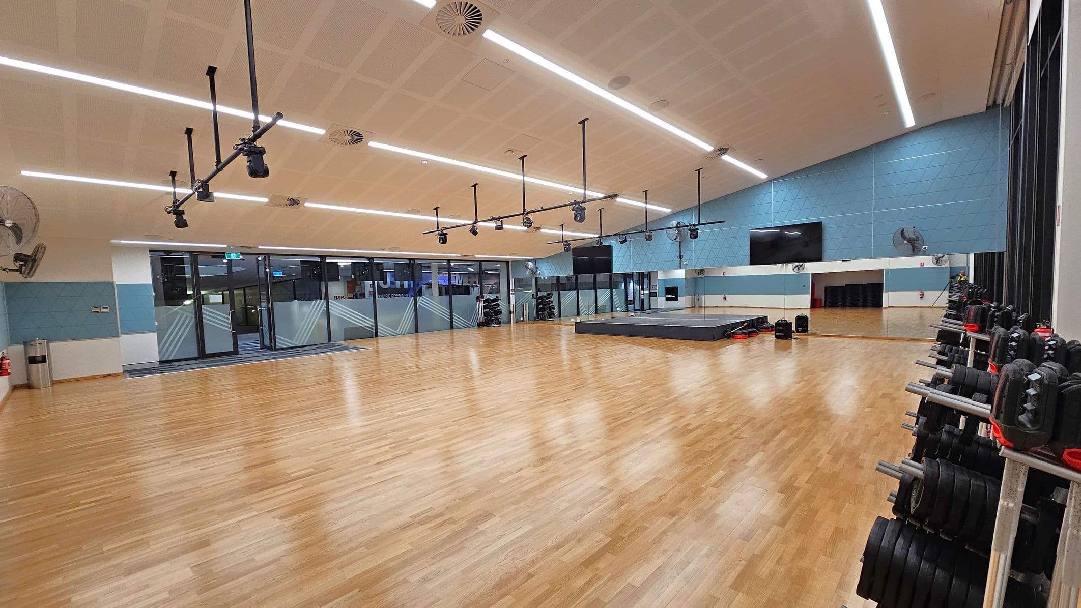 A spacious gym studio with a wooden floor and a high ceiling featuring linear lights. On the right, there are racks of weights neatly organised. The back wall is light blue with a large mirror and a mounted television. In the centre, a raised platform is visible. The left side has large glass doors and windows with geometric patterns, leading to another area. Ceiling-mounted speakers and fans are distributed throughout the room.