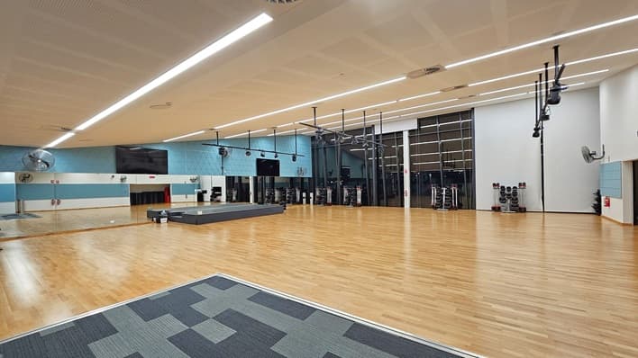 A spacious gym studio with a wooden floor and a patterned carpet section in the foreground. The left wall features large mirrors and a mounted TV, with blue panels above. Ceiling fans and lighting fixtures are suspended from the ceiling. The right side has a white wall with exercise equipment, including weights and step platforms. The back wall is mostly glass, allowing natural light, and more equipment is visible. The ceiling is lined with long fluorescent lights.