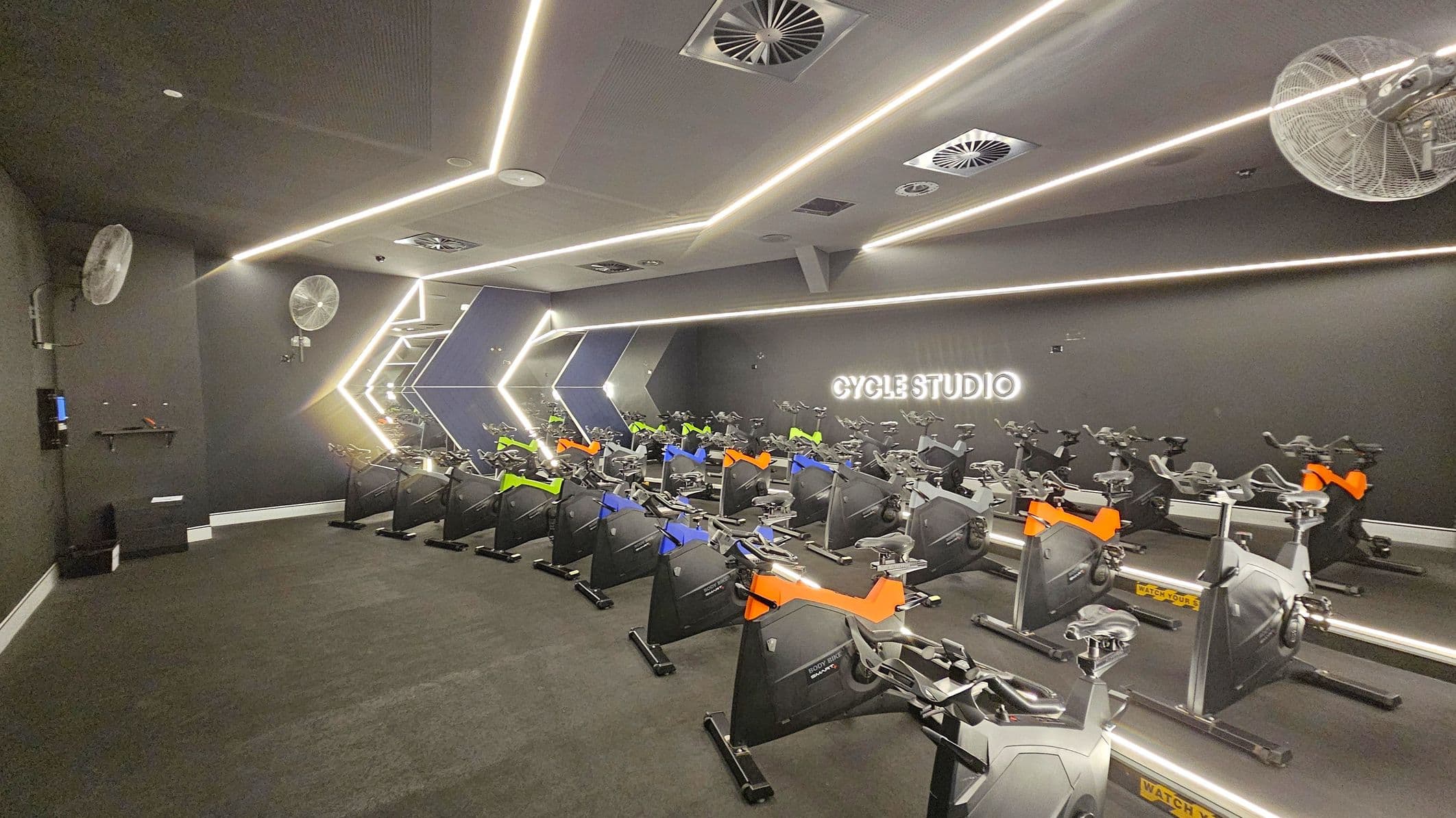 A modern cycling studio with a dark interior. On the right, a neon sign reads "CYCLE STUDIO" on the wall. The room is filled with stationary bikes, each with colourful accents in orange, blue, and green. The ceiling features linear LED lights and ventilation units. Fans are mounted on the walls. The floor is carpeted in dark grey, and the walls have geometric light patterns.