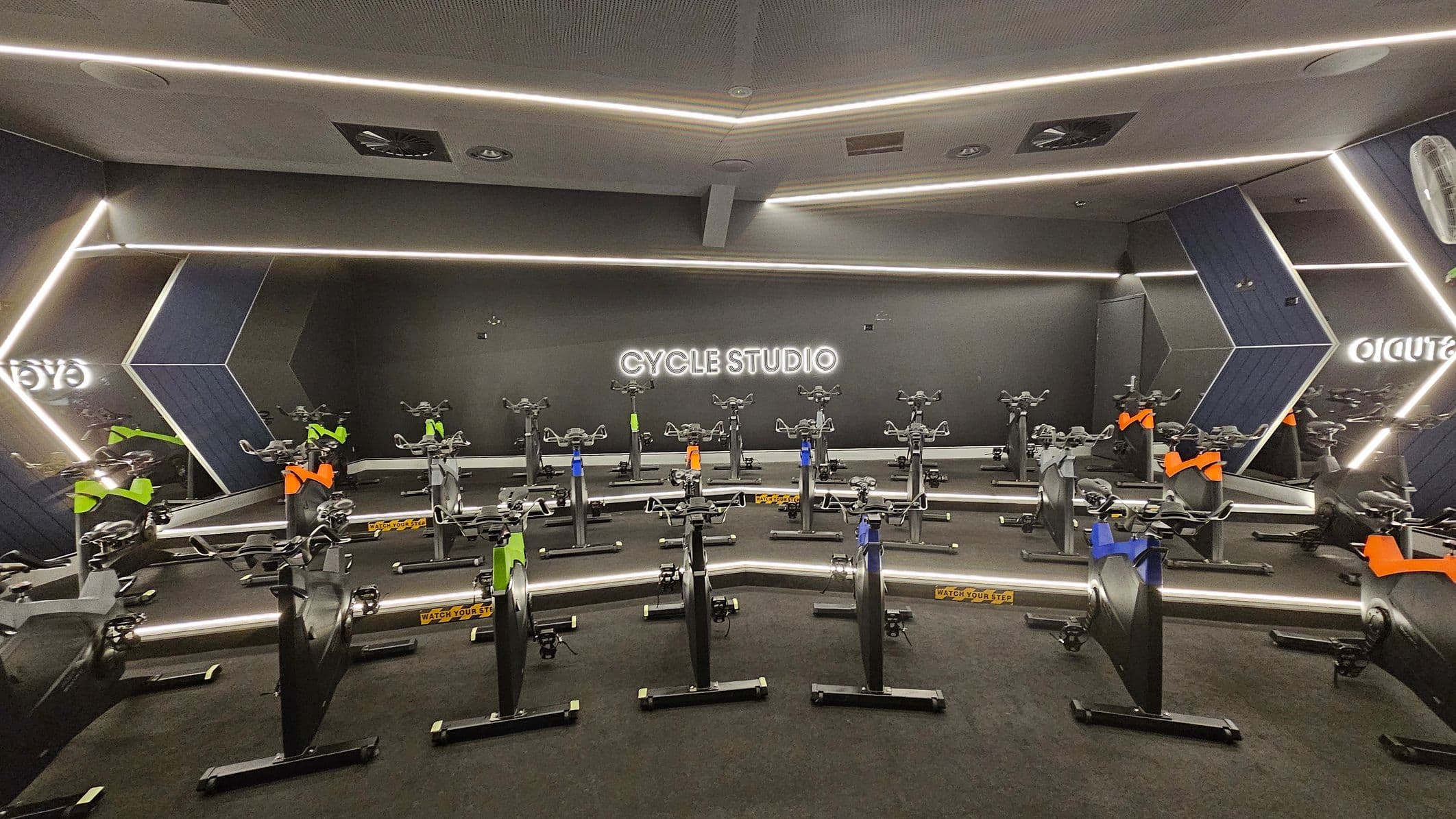 A modern cycle studio with a dark interior. In the centre, several stationary bikes are arranged in rows, each with colourful accents like green, blue, and orange. The back wall features a neon sign reading "CYCLE STUDIO". The walls have geometric patterns with reflective surfaces, and the ceiling has recessed lighting and ventilation panels. The floor is carpeted in dark material, and there are caution signs reading "WATCH YOUR STEP".
