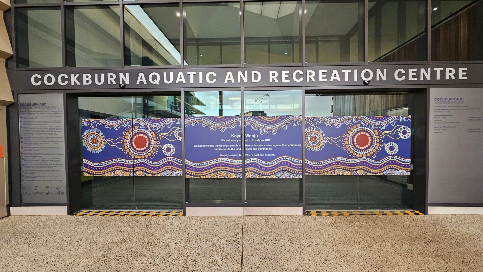 Entrance to the Cockburn Aquatic and Recreation Centre with large glass doors featuring vibrant Indigenous artwork. The artwork includes circular patterns and wavy lines in red, yellow, white, and blue. Above the doors, the centre's name is displayed in bold white letters. On the left, a panel lists conditions of entry. The floor is speckled concrete, and the ceiling is not visible.