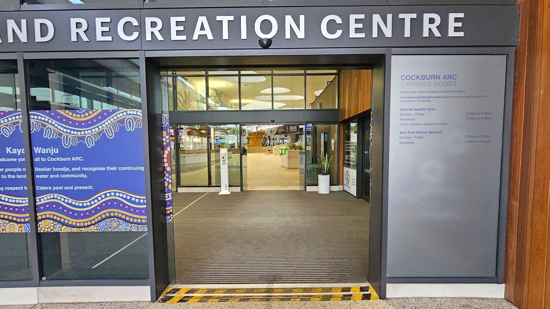 Entrance to a recreation centre with large glass doors. Above the entrance, "COCKBURN AQUATIC AND RECREATION CENTRE" is displayed in bold letters. To the left, a blue panel with decorative patterns and text. To the right, a grey panel lists opening hours. Inside, a spacious lobby with a staircase, potted plants, and various signs. The floor is carpeted, and the ceiling features circular lights.