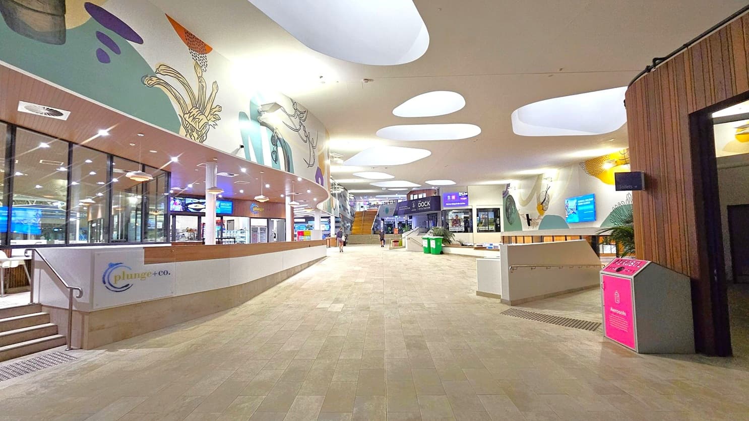 A spacious indoor area with a light-coloured tiled floor. On the left, there's a café named "plunge+co." with glass windows and a counter. The ceiling features large, irregularly shaped cut-outs allowing light through. The walls are decorated with abstract art. In the centre, a wide walkway leads to a staircase. On the right, there's a pink recycling bin and a sign for toilets. The area is well-lit with overhead lights and has a modern design.