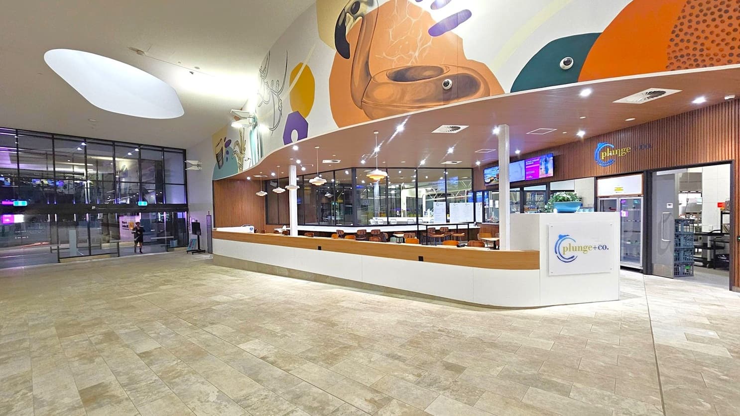 A spacious indoor area with a modern design. On the right, there's a café counter with a sign reading "plunge+co." The counter is white with wooden accents, and several pendant lights hang above. Behind the counter, there are tables and chairs. The left side features large glass doors leading outside. The ceiling is high with a large, oval-shaped light fixture. The floor is tiled in a light, neutral colour. A colourful mural decorates the upper wall above the café.