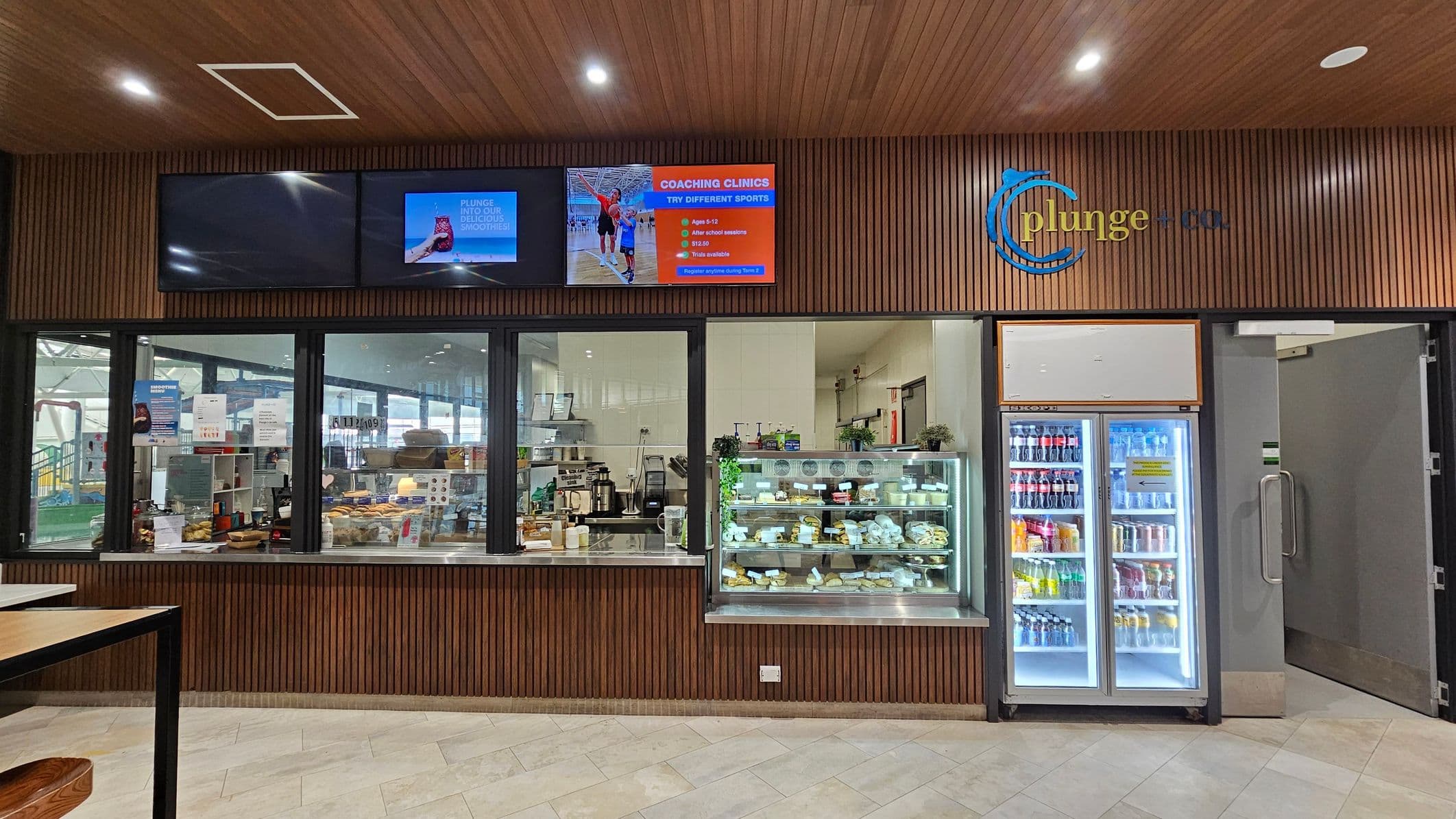 A café counter with a wooden panelled wall and ceiling. On the left, there are two digital screens displaying advertisements. Below, a glass window shows the café interior with various items and equipment. In the centre, a display case is filled with pastries and snacks. On the right, a refrigerator stocked with assorted drinks stands next to an open door. The floor is tiled in a light colour.