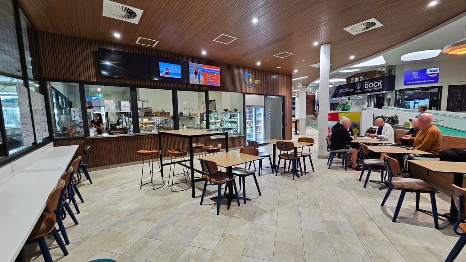 Interior of a café with a wooden ceiling and light-coloured tiled floor. On the left, a counter with high stools and a window showing a staff member inside. In the centre, several wooden tables and chairs are arranged. On the right, a group of people sit at a table, engaged in conversation. The background features a refrigerated display and signage for "The Dock" and "Plunge." Bright lighting and a modern design create an inviting atmosphere.