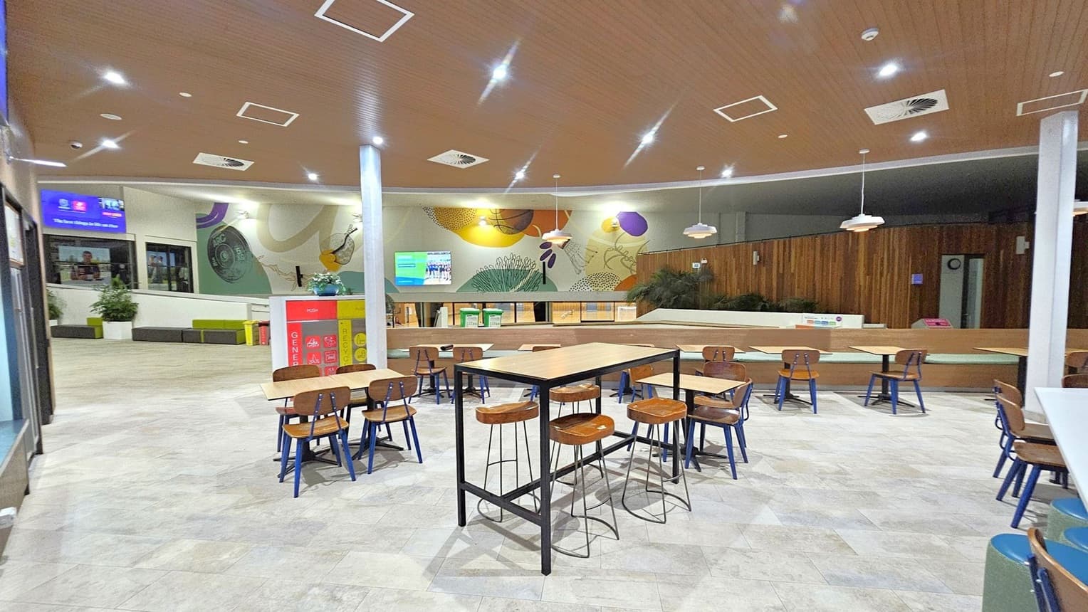 A spacious indoor area with a light-coloured tiled floor and a wooden ceiling with recessed lighting. In the centre, there are tables and chairs, including a high table with stools. The background features a mural with abstract designs and a wall-mounted screen. To the left, there are windows and potted plants. The right side has wooden panelling and more seating. Hanging lights are evenly spaced across the ceiling.