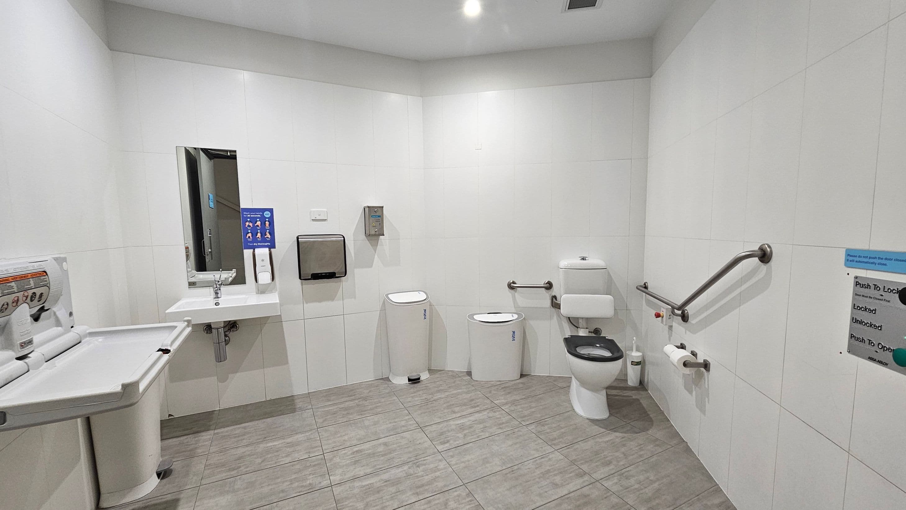 A spacious, accessible public toilet with white tiled walls and a grey tiled floor. On the left, a baby changing station is mounted on the wall. In the centre, a sink with a mirror above it is accompanied by a soap dispenser and a paper towel holder. To the right, a toilet with a black seat is equipped with grab bars for accessibility. A waste bin and a sanitary bin are nearby. A sign with instructions is visible on the right wall. The ceiling has recessed lighting.