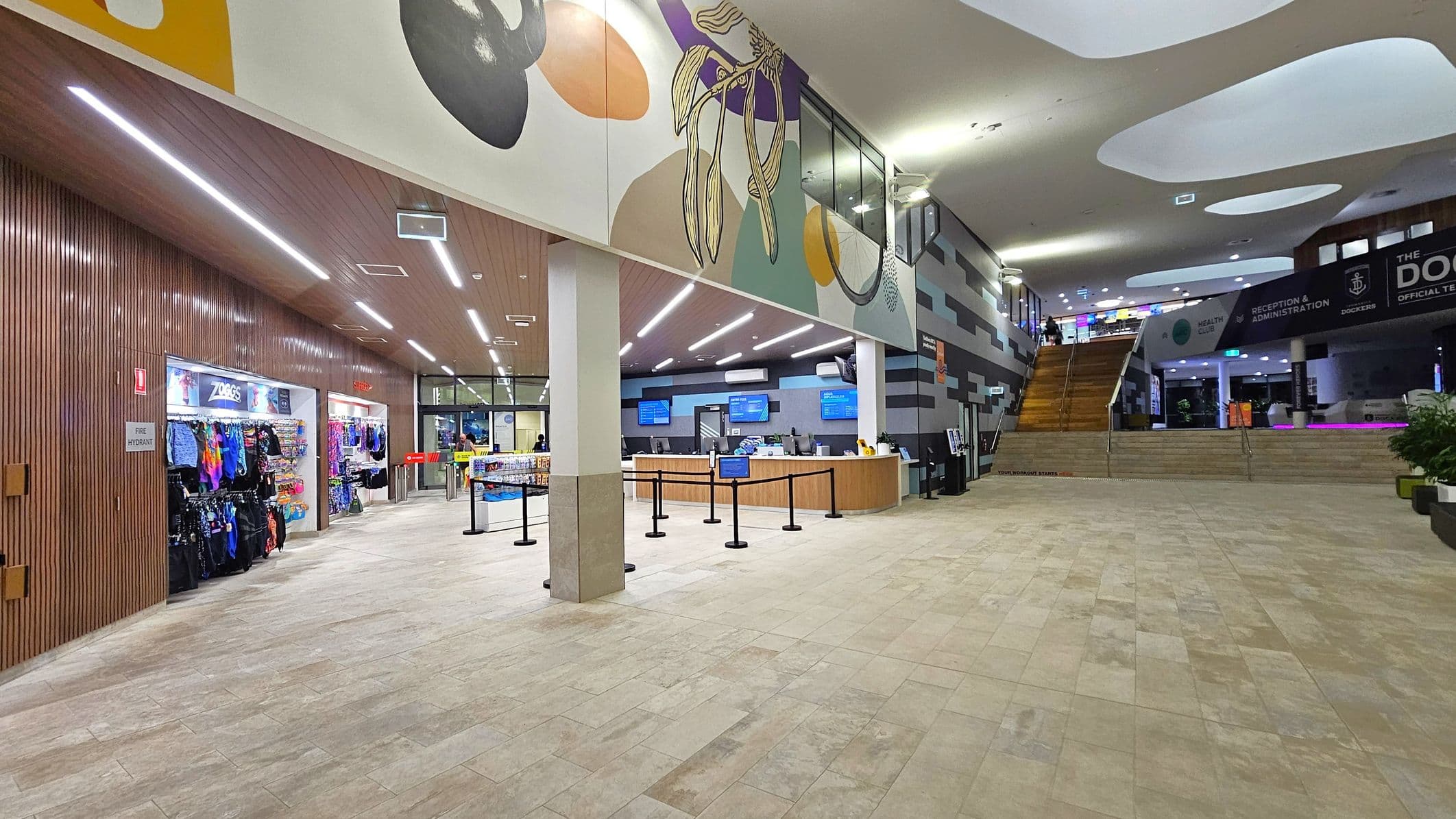 A spacious indoor area with a tiled floor. On the left, there is a shop displaying colourful swimwear and accessories. In the centre, a reception desk with queue barriers is visible, backed by digital screens. The ceiling features artistic designs with abstract patterns. On the right, a staircase leads to an upper level with signs for health and administration. The walls have a mix of wooden and patterned designs, and there are potted plants near the stairs.