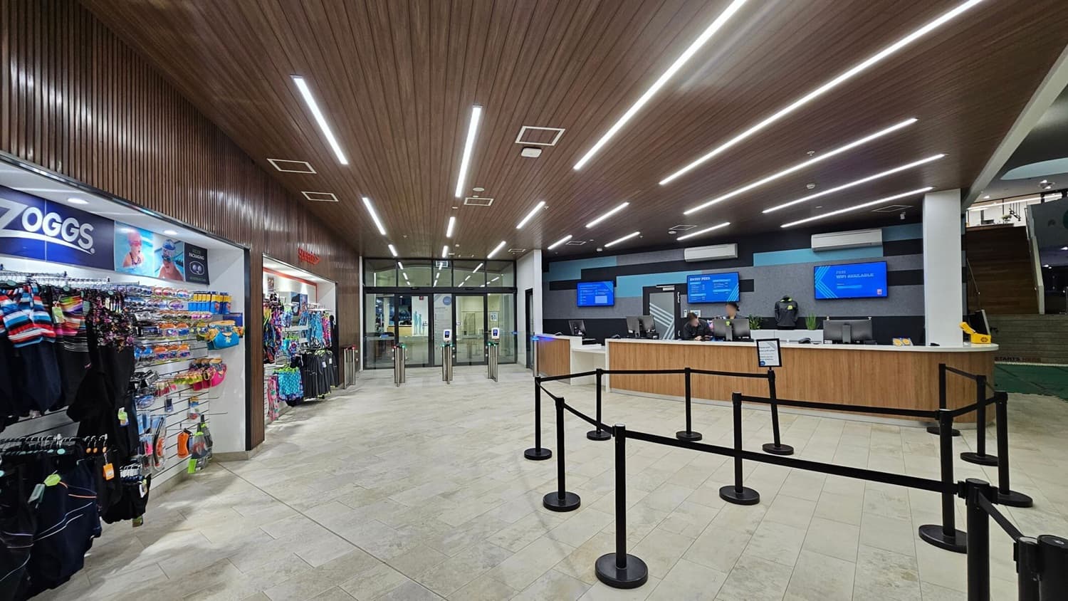 A spacious indoor area with a shop on the left displaying swimwear and accessories. In the centre, there are turnstiles leading to glass doors. On the right, a reception desk with two staff members and digital screens above displaying information. The floor is tiled in light grey, and the ceiling features wooden panelling with linear lights. Black queue barriers are set up in front of the desk.