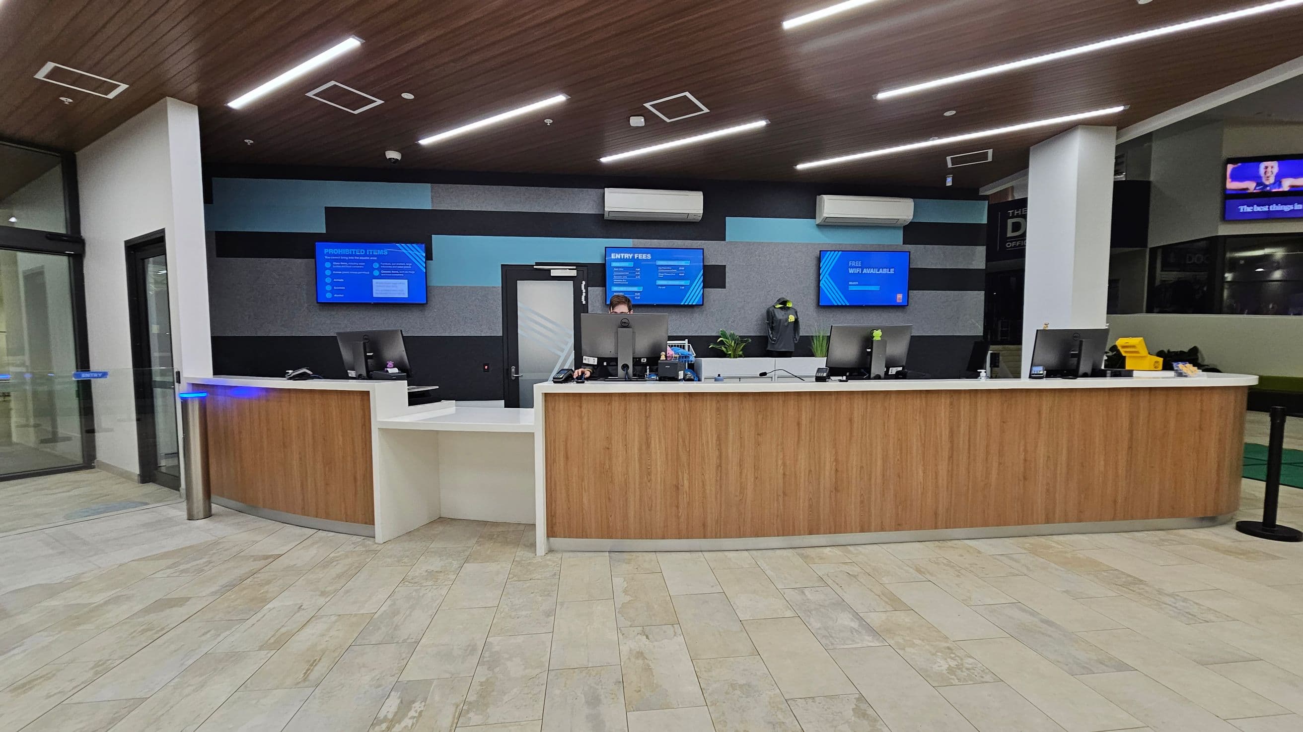 A modern reception area with a wooden front desk and multiple computer monitors. Behind the desk, three wall-mounted screens display information. The wall features a geometric pattern in shades of blue and grey. There are air conditioning units above the screens. The floor is tiled in a light, mixed pattern, and the ceiling is wooden with recessed lighting. A glass door is on the left, and a small plant is visible on the desk.