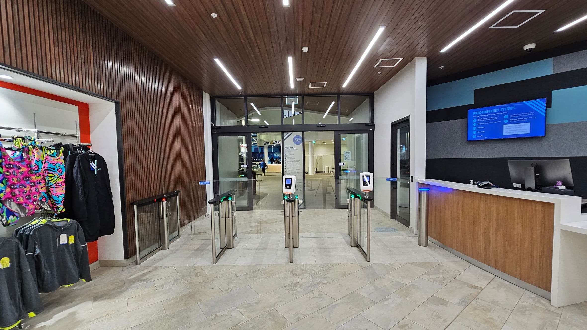 On the left, there is a display of colourful clothing, including bright pink and patterned items, hanging on racks. In the centre, there are turnstiles with digital screens, leading to glass doors. The floor is tiled in light colours. On the right, a reception desk with a computer and a blue information screen above it. The ceiling is wooden with linear lights.