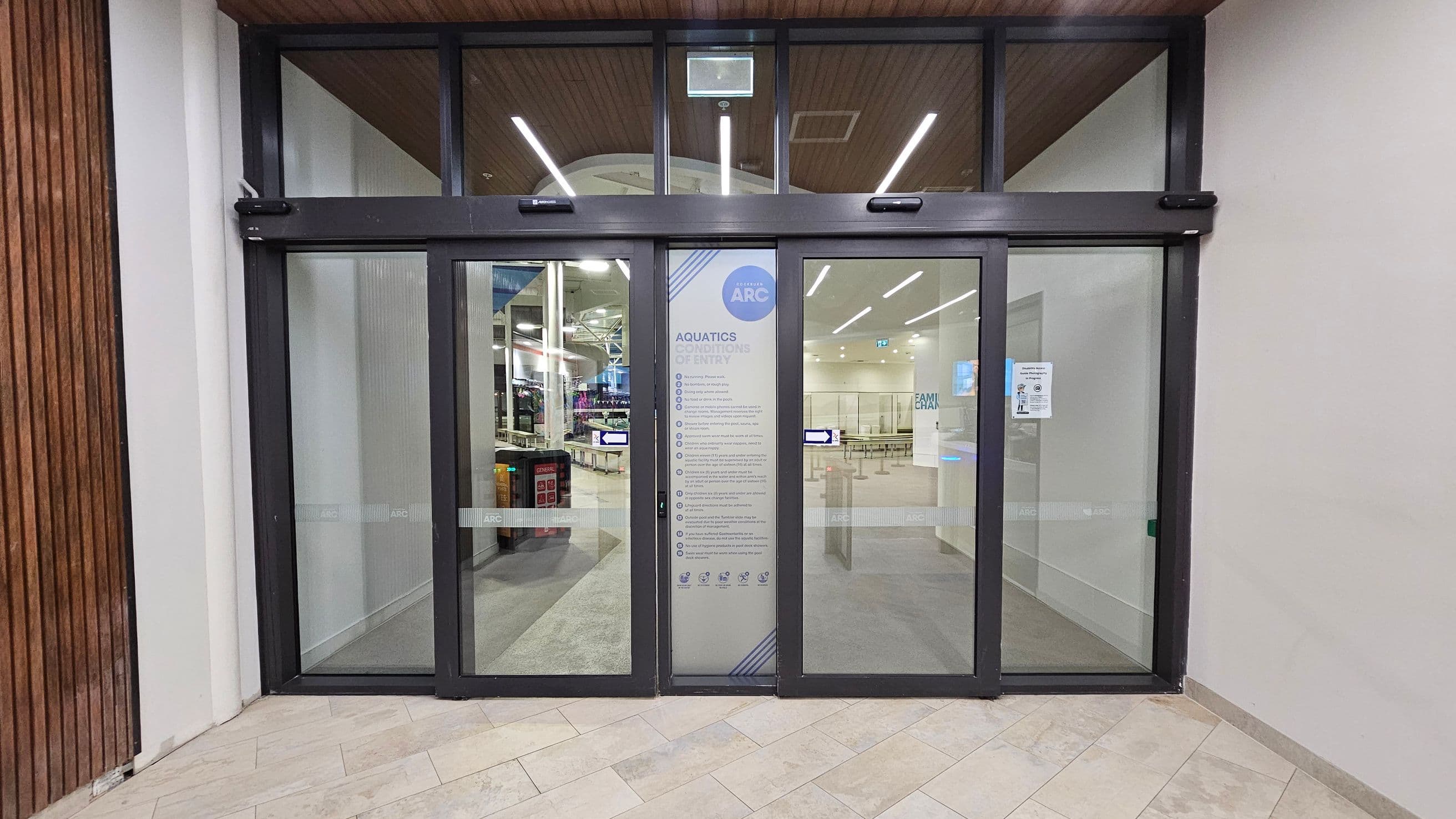 Glass entrance with automatic sliding doors, featuring a central panel displaying "Aquatics Conditions of Entry" and a logo. Inside, a spacious area with visible gym equipment and a reception desk. The ceiling has wooden panels and linear lights. The floor outside is tiled in a light beige pattern, while the interior floor is grey. Walls are white, with a wooden panel on the left side.