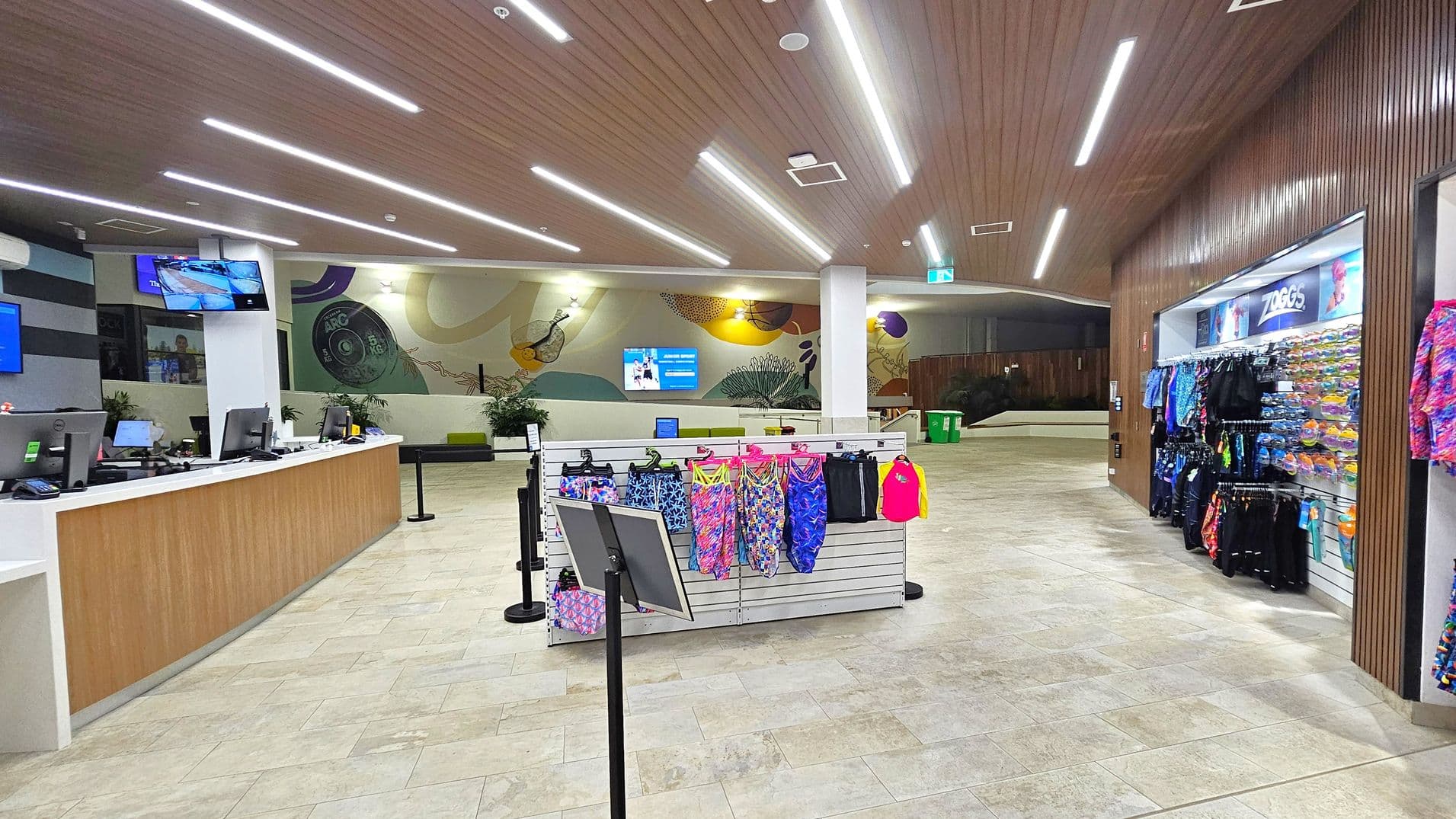A spacious indoor area with a tiled floor and a wooden ceiling featuring linear lights. On the left, there's a long reception desk with computers and monitors. In the centre, a display rack holds colourful swimwear. The right side has a wall-mounted display of more swimwear and accessories. The background features a mural with abstract designs and a seating area with green benches. A digital screen is mounted on the wall, and a green bin is visible in the distance.