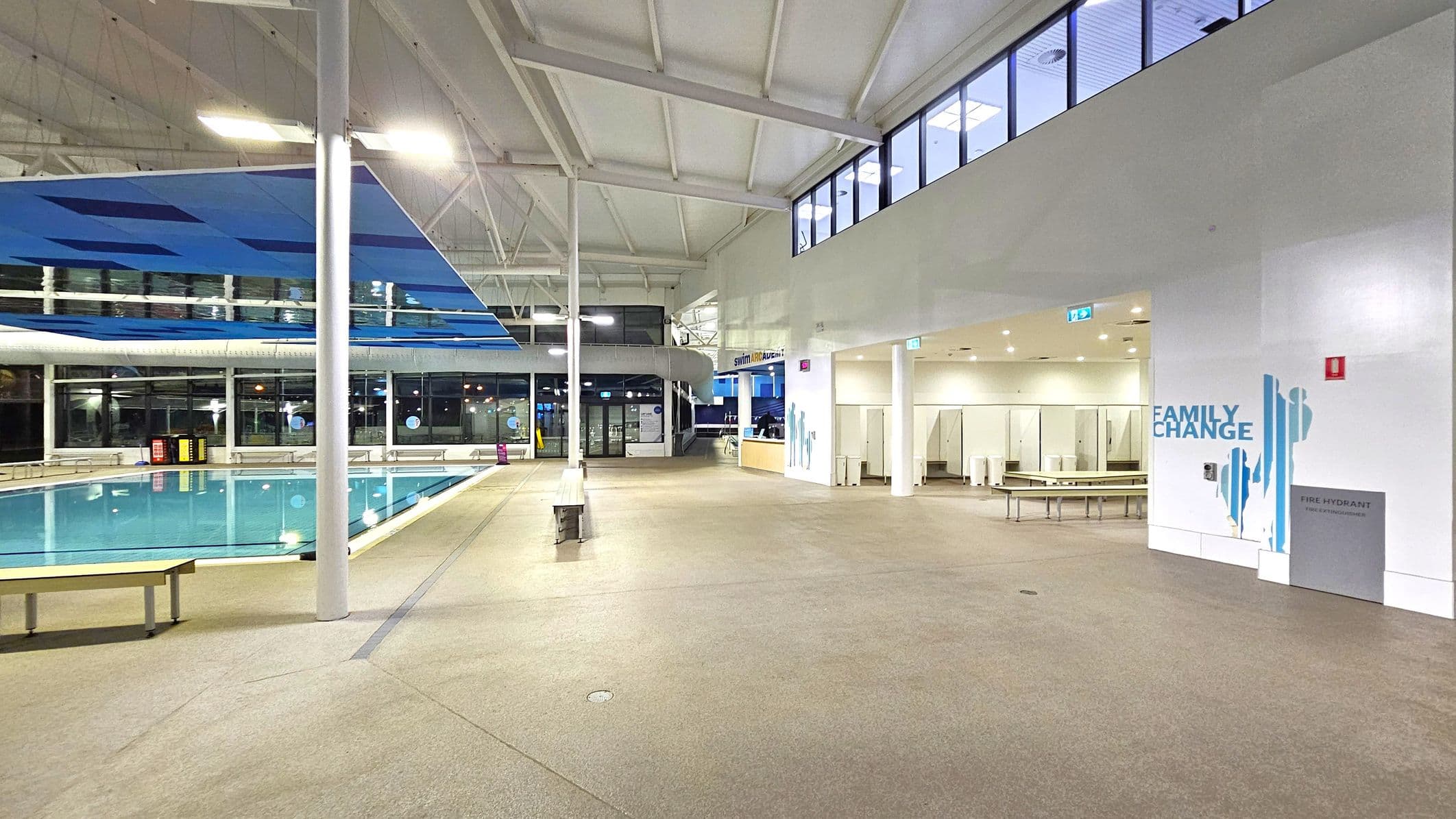 Indoor swimming pool area with a large pool on the left, surrounded by glass windows. Above the pool, blue panels hang from the ceiling. On the right, there's a "Family Change" area with benches and changing cubicles. The floor is a light brown, non-slip surface. The ceiling is high with exposed beams and lights. A sign for a swim academy is visible near the entrance to the changing area.