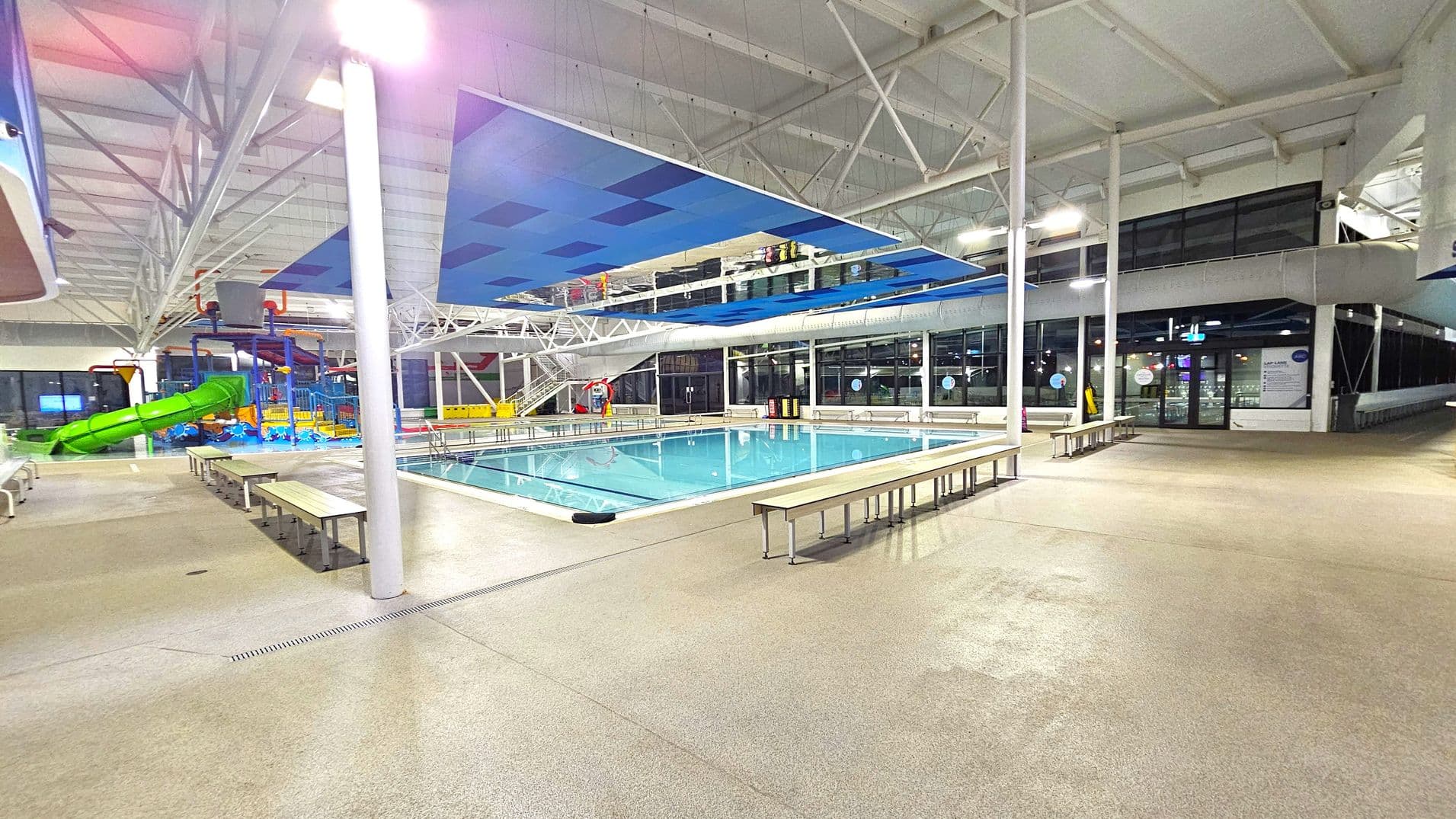 Indoor swimming pool area with a large rectangular pool in the centre. Surrounding the pool are several benches on a beige tiled floor. On the left, a colourful water play area with a green slide is visible. The ceiling is high with exposed beams and blue acoustic panels. Large windows line the right side, allowing natural light to enter. The area is well-lit with overhead lights.