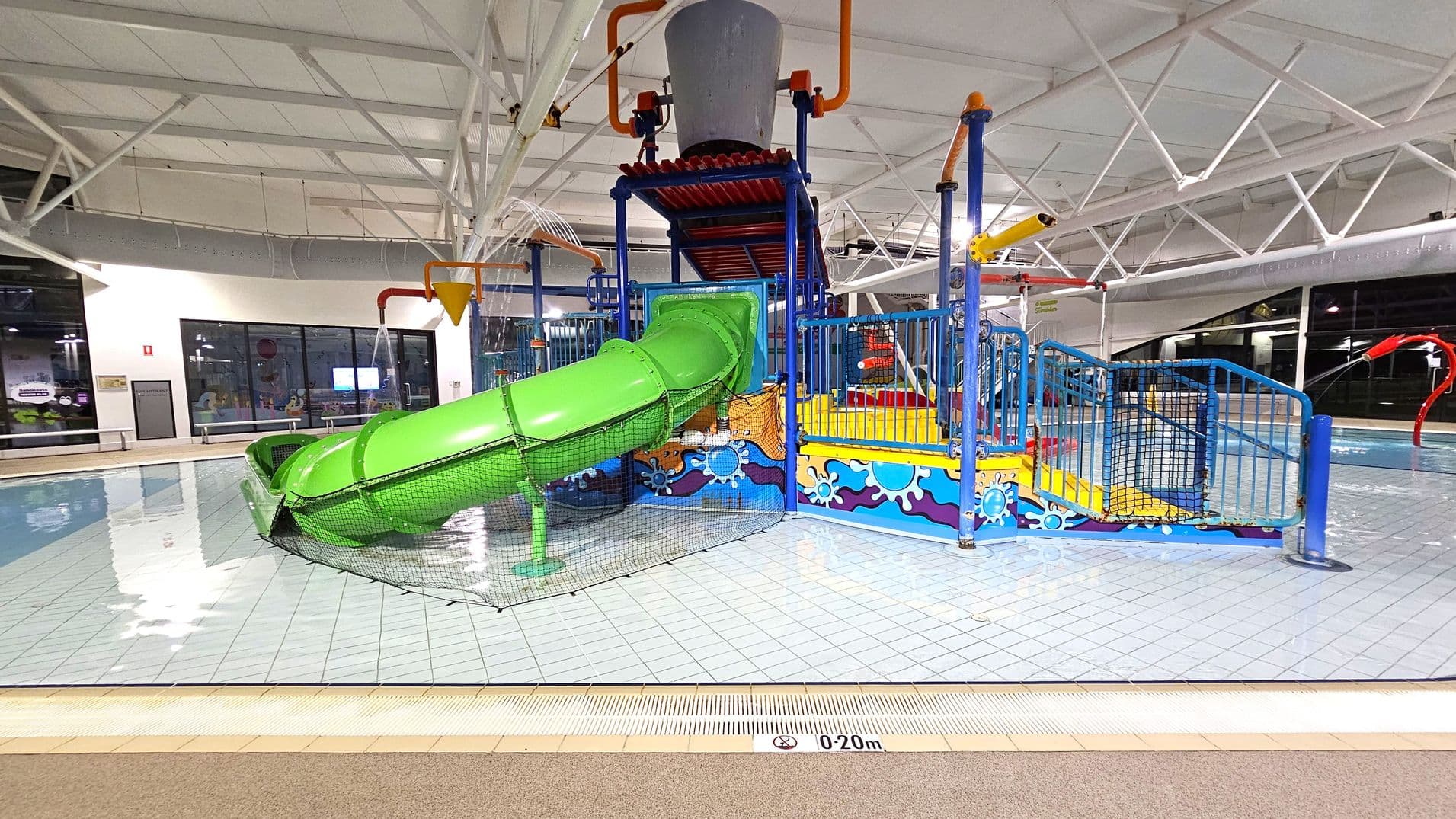 A colourful indoor water play area with a bright green slide on the left, leading into a shallow pool. The structure is blue and yellow with decorative patterns, featuring a large grey bucket on top. Water sprays from various points, and there are orange and red pipes. The floor is tiled, and the ceiling has white beams. Large windows are visible in the background, and a sign indicates a depth of 0-20 cm.