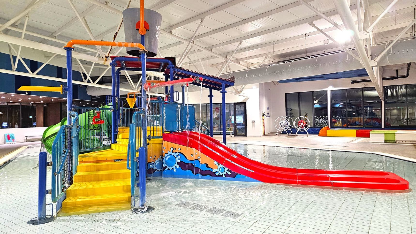 Indoor water play area with a colourful structure featuring yellow steps, a red slide, and blue support beams. A large grey bucket is positioned above, with water pouring from it. The floor is tiled and wet, with shallow water surrounding the play equipment. In the background, there are large windows and a wall with colourful accents. The ceiling is high with white beams and lighting fixtures.
