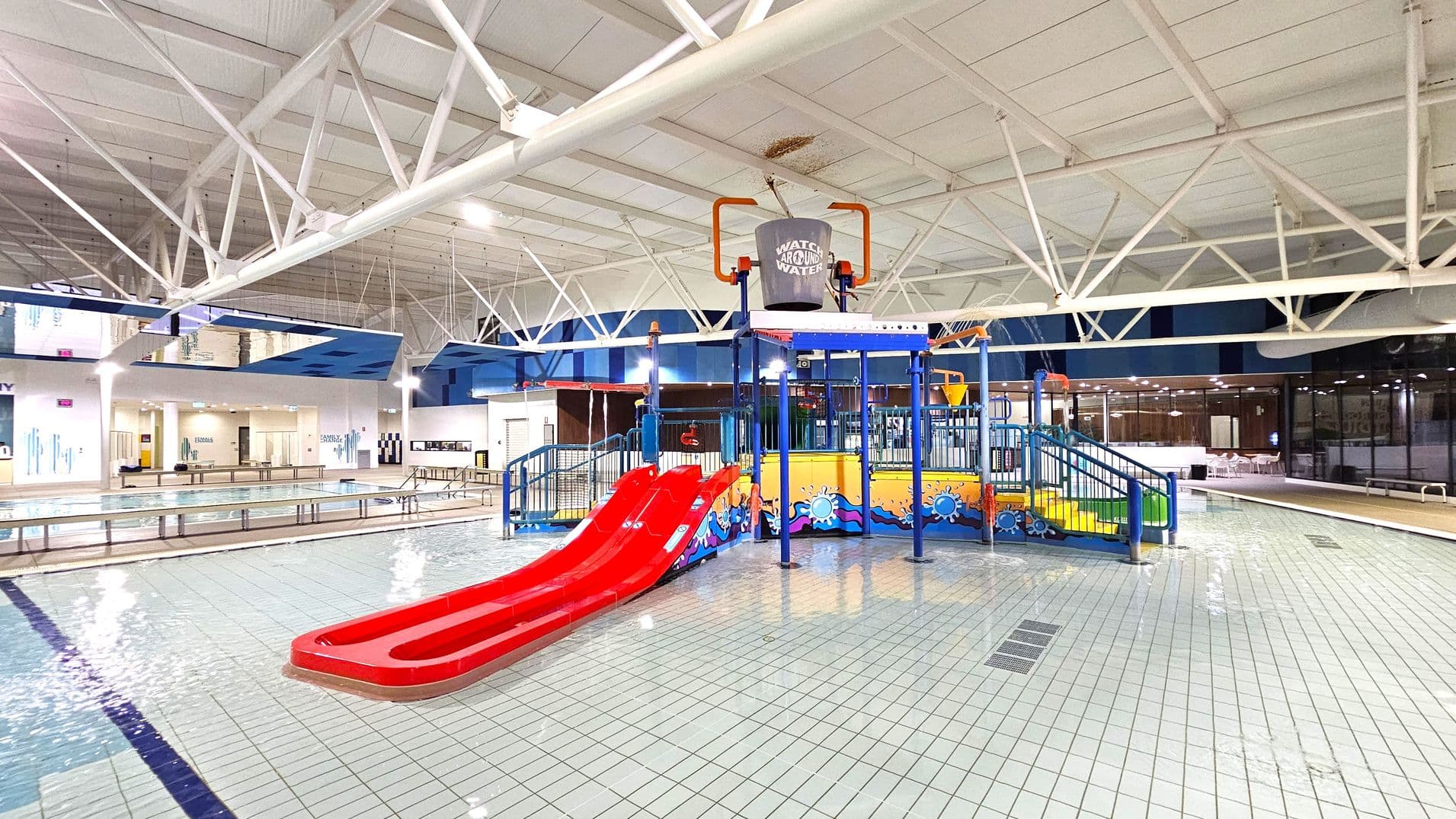 Indoor swimming pool area with a children's play structure in the centre. The structure features a red double slide, blue railings, and a large bucket on top. The floor is tiled in white with blue lines. To the left, there are lanes for swimming, and to the right, a seating area with glass walls. The ceiling is high with white beams and lighting fixtures. The background includes a wall with doors and signage.