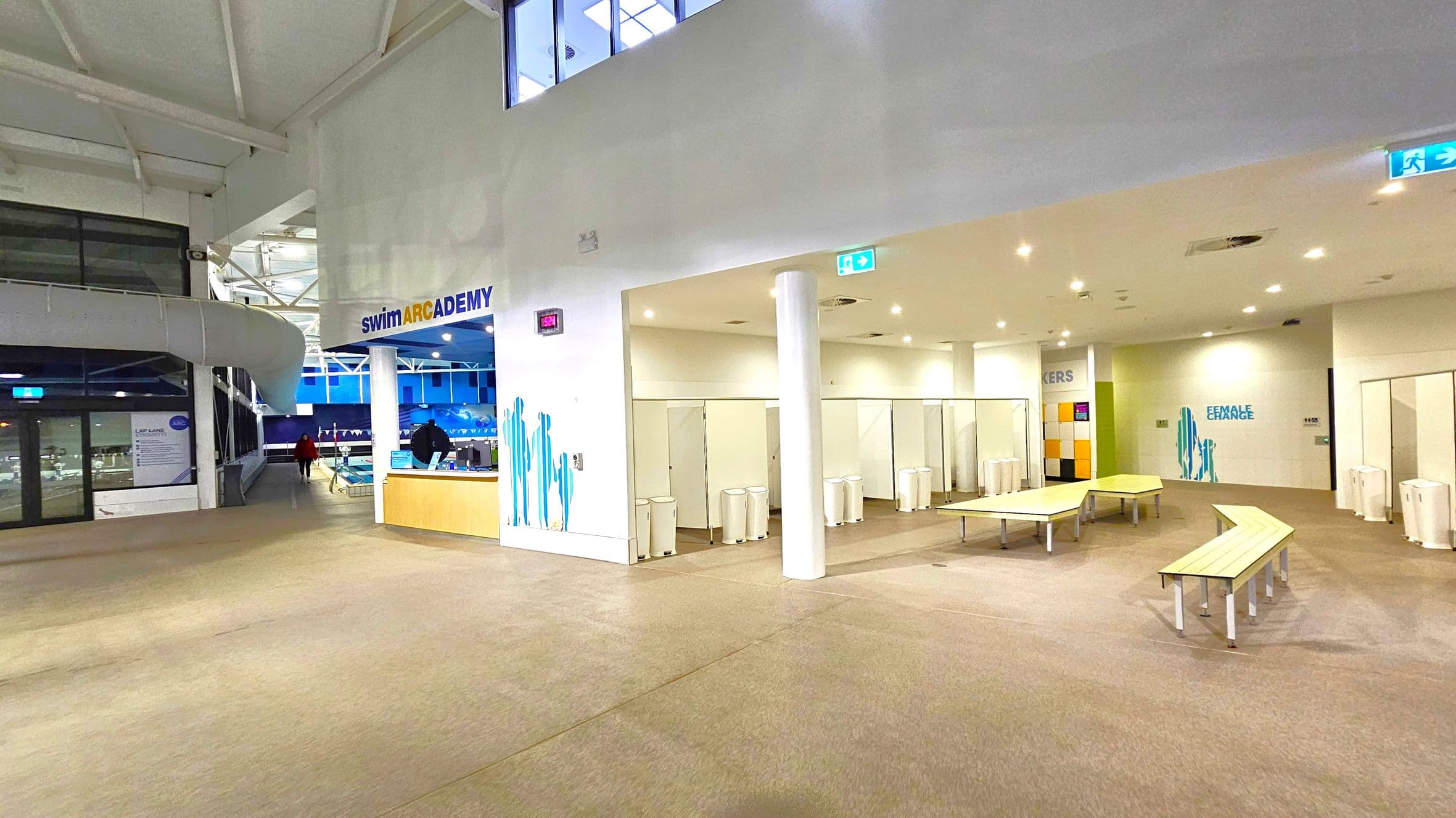 A spacious indoor area with a high ceiling. On the left, there's a reception desk with a sign reading "swimARCADEMY" above it. Behind the desk, a pool area is visible through large windows. On the right, there are changing rooms with white doors and several white bins. Benches are arranged in a zigzag pattern in front of the changing rooms. The floor is carpeted, and the ceiling has recessed lighting. Signs indicate "LOCKERS" and "FEMALE CHANGE" areas.