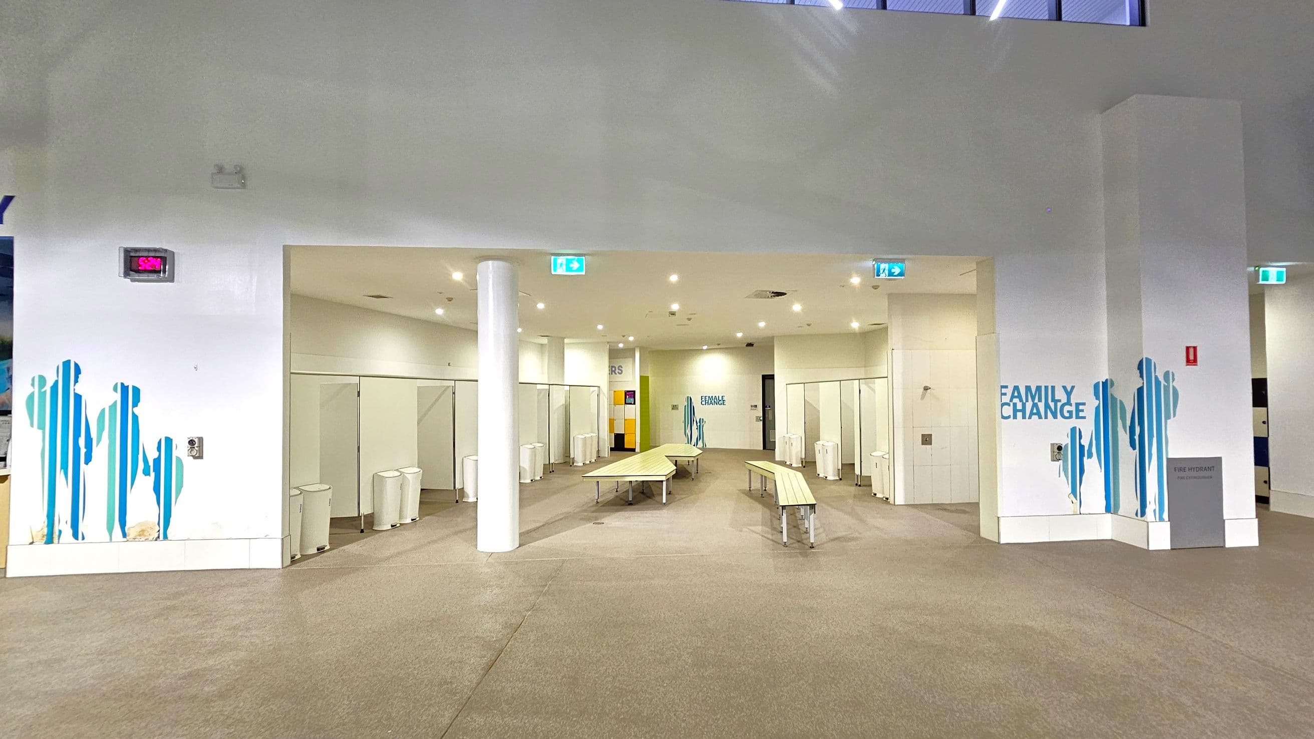 A spacious changing area with a beige carpeted floor and a high white ceiling. In the centre, there are several long benches. On the left and right, there are rows of changing cubicles with white doors. Blue silhouettes of people are painted on the walls, and "FAMILY CHANGE" is written on the right wall. A digital clock displays the time as 5:24. Emergency exit signs are visible above the entrances.