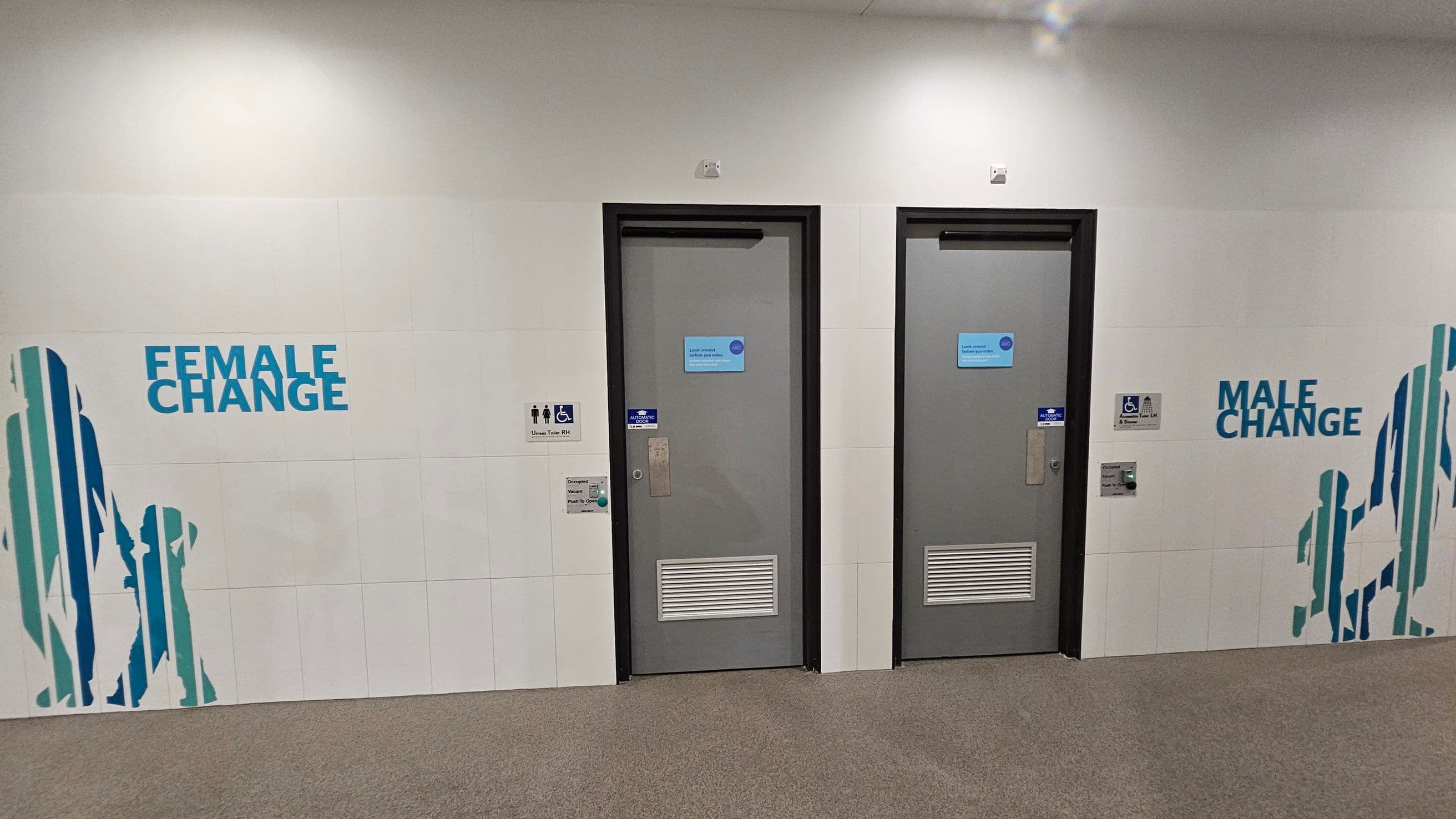 Two grey doors are centred on a white tiled wall. The left door is labelled "Female Change" and the right "Male Change". Blue silhouettes of people are painted on the wall near each door. Between the doors, there are signs indicating accessible facilities. The floor is covered with brown carpet, and the ceiling is plain white.