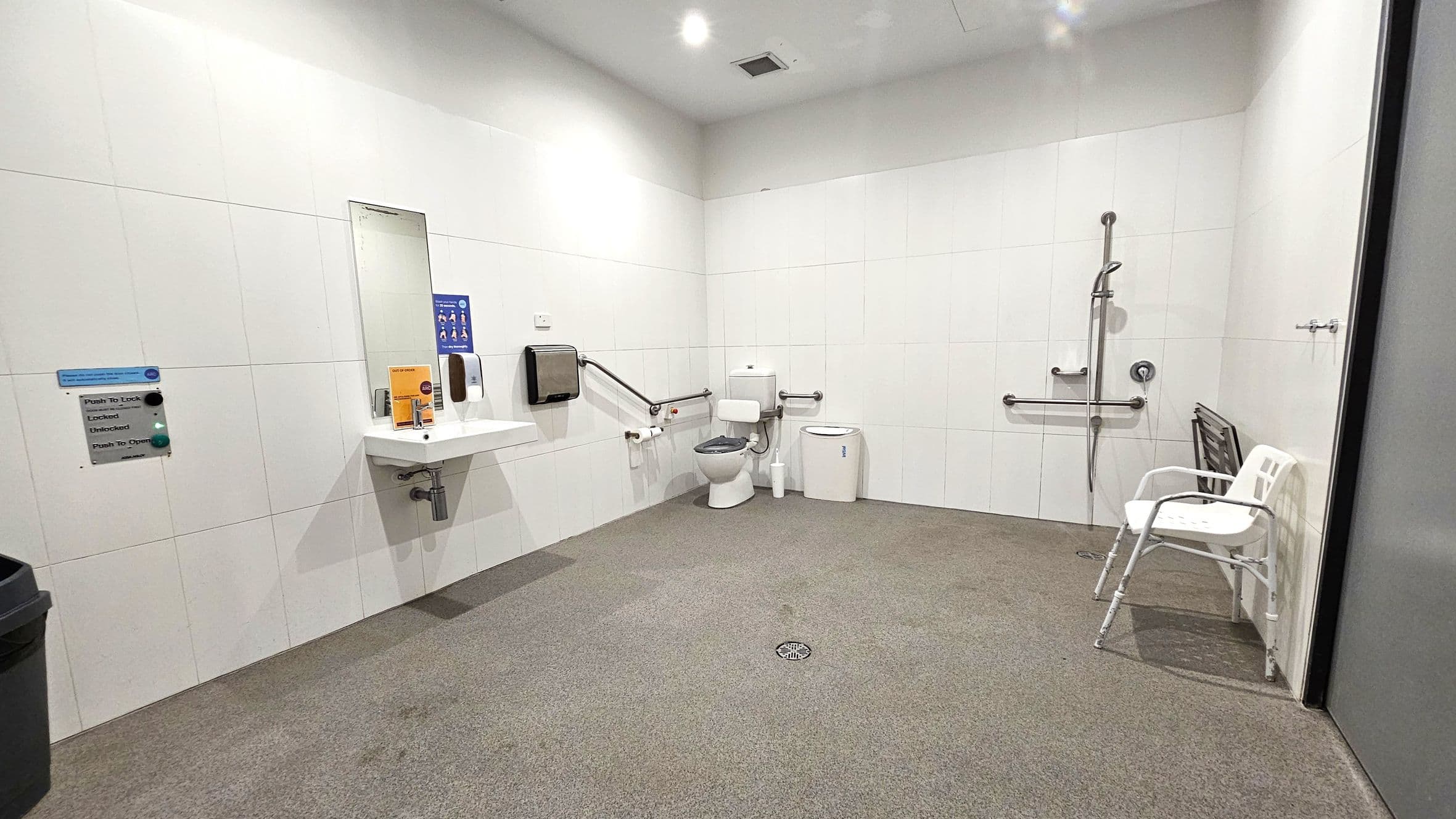 A spacious, accessible bathroom with white tiled walls and a speckled floor. On the left, a sink with a mirror above it and a soap dispenser. A hand dryer is mounted nearby. In the centre, a toilet with support rails and a sanitary bin. On the right, a shower area with a handheld showerhead and additional support rails. A white plastic chair is placed against the wall. The ceiling is plain with recessed lighting.