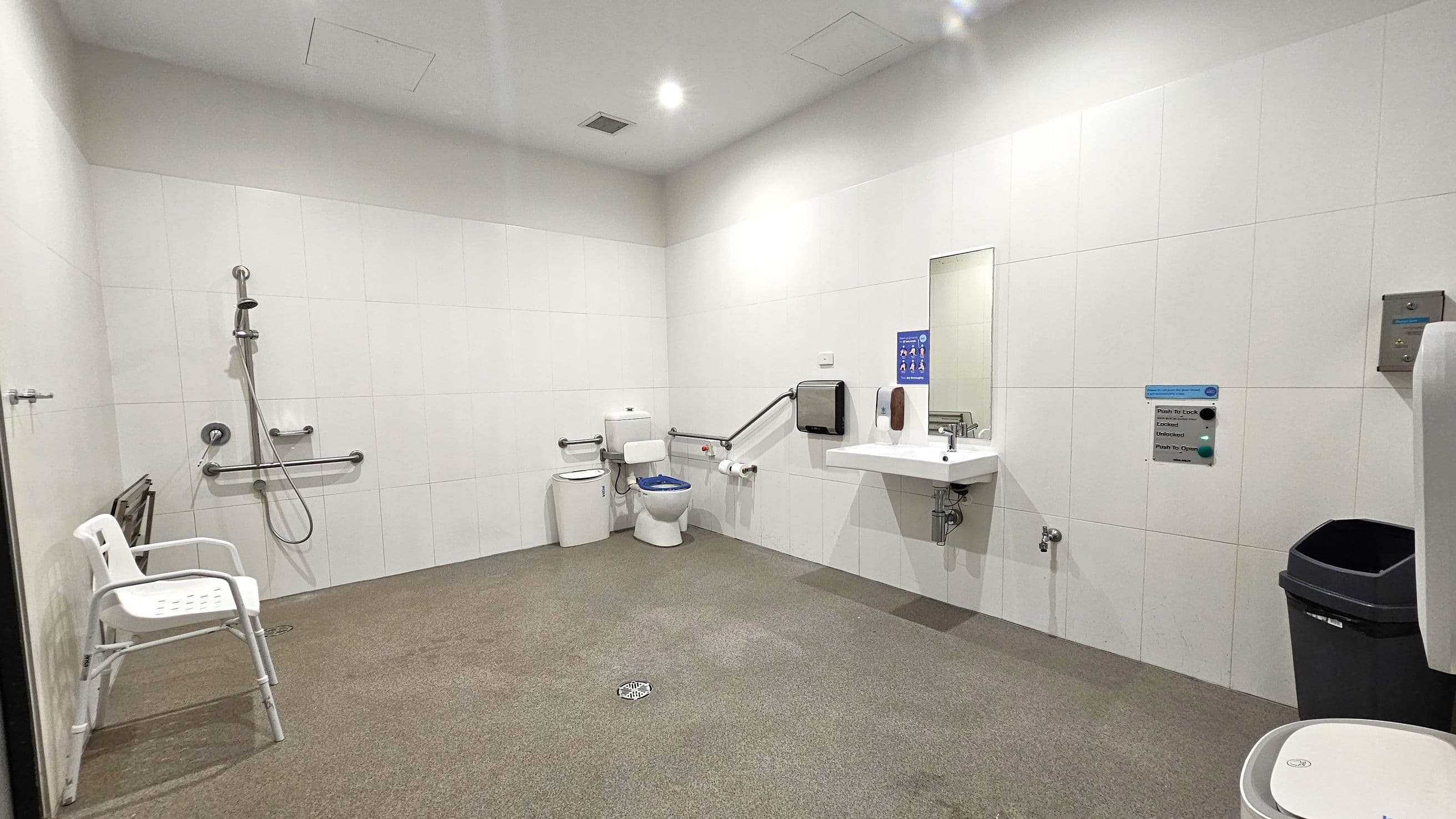 A spacious, accessible bathroom with white tiled walls and a speckled floor. On the left, a shower area with a handheld showerhead and grab bars, accompanied by a white plastic chair. In the centre, a toilet with support rails and a blue seat cover. To the right, a sink with a mirror above it, a soap dispenser, and a paper towel dispenser. A black bin is positioned near the sink. The ceiling is white with recessed lighting.