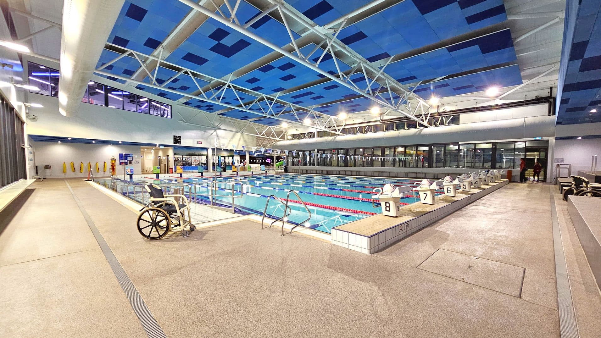 Indoor swimming pool with multiple lanes, each marked with a number. On the left, a wheelchair is positioned near the poolside. The ceiling features a blue and white geometric pattern with exposed beams. The right side has a row of starting blocks numbered 1 to 8. In the background, large windows and a wall with life vests hanging. The floor is a light grey, non-slip surface.