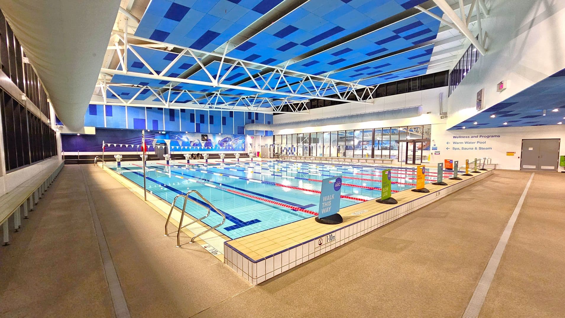 Indoor swimming pool with multiple lanes, bordered by a tiled edge. On the left, a row of benches lines the wall beneath large windows. The right side features signs indicating different lanes and directions to wellness areas. The ceiling is blue with white structural beams, and the floor is a light-coloured non-slip surface. In the background, a wall with blue panels and a mural, along with glass doors leading to other facilities.