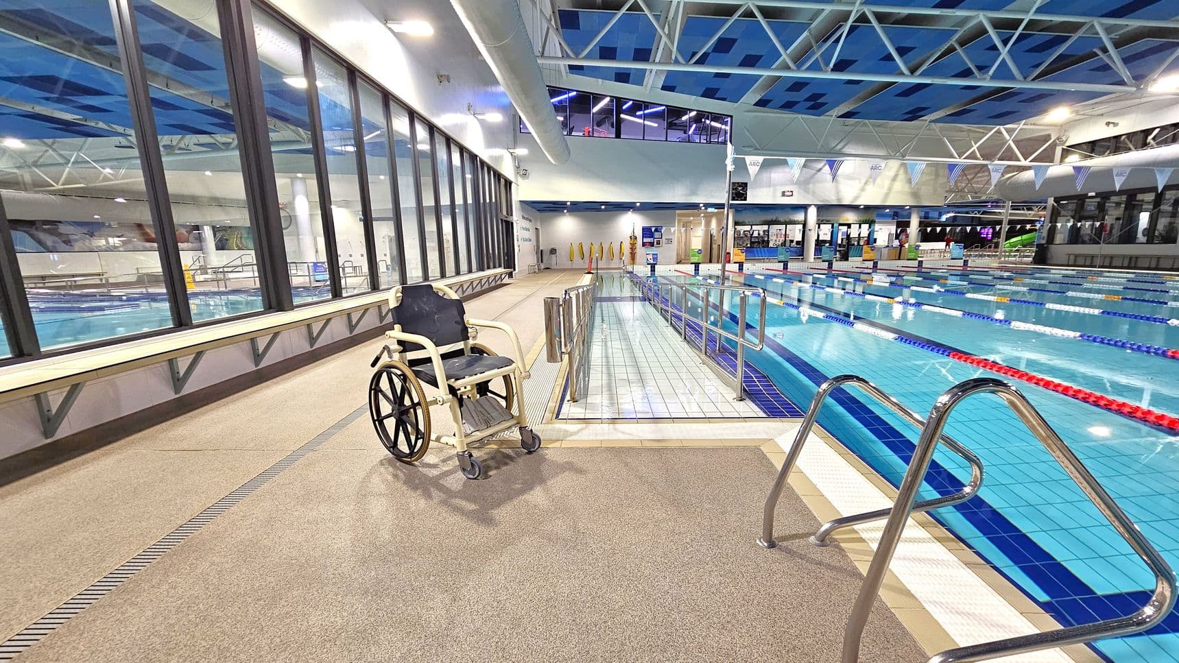A swimming pool area with a wheelchair on the left side, positioned on a beige tiled floor. Large windows run along the left wall, providing a view of another pool. In the centre, a ramp with metal railings leads into the pool, which has marked lanes with red, blue, and yellow lane dividers. The ceiling features a blue and white geometric pattern with exposed beams. Bright lighting illuminates the space, and there are various signs and equipment visible in the background.