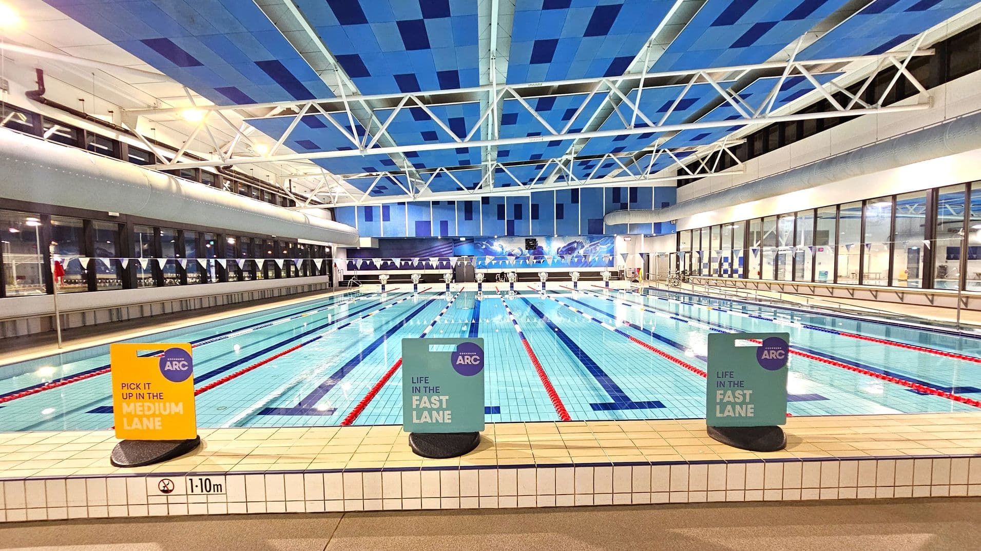 Indoor swimming pool with multiple lanes, each marked by red and blue lane dividers. The pool is surrounded by a tiled deck with signs indicating lane speeds. The ceiling features a geometric pattern of blue panels and white structural beams. Large windows line the left and right walls, allowing natural light. The pool's depth is marked as 1.10 metres. The background shows a mural with aquatic imagery.