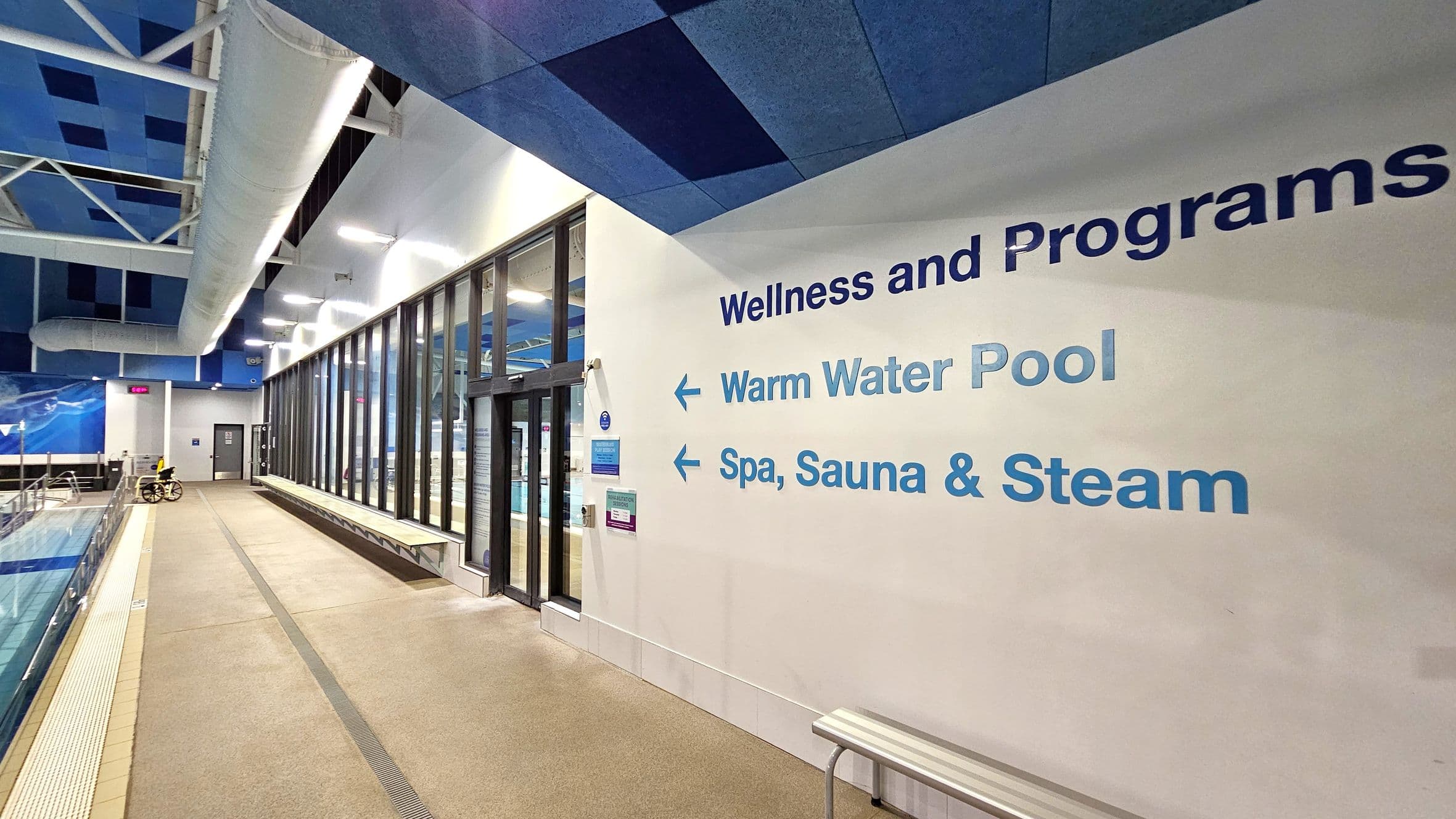 A swimming pool area with a beige tiled floor and a blue and white ceiling featuring large ducts. On the right, a wall displays the words "Wellness and Programs" with arrows pointing left to "Warm Water Pool" and "Spa, Sauna & Steam." Large glass windows and doors line the wall, reflecting the pool area. A yellow wheelchair is visible near the pool on the left, and a bench is positioned against the wall on the right.