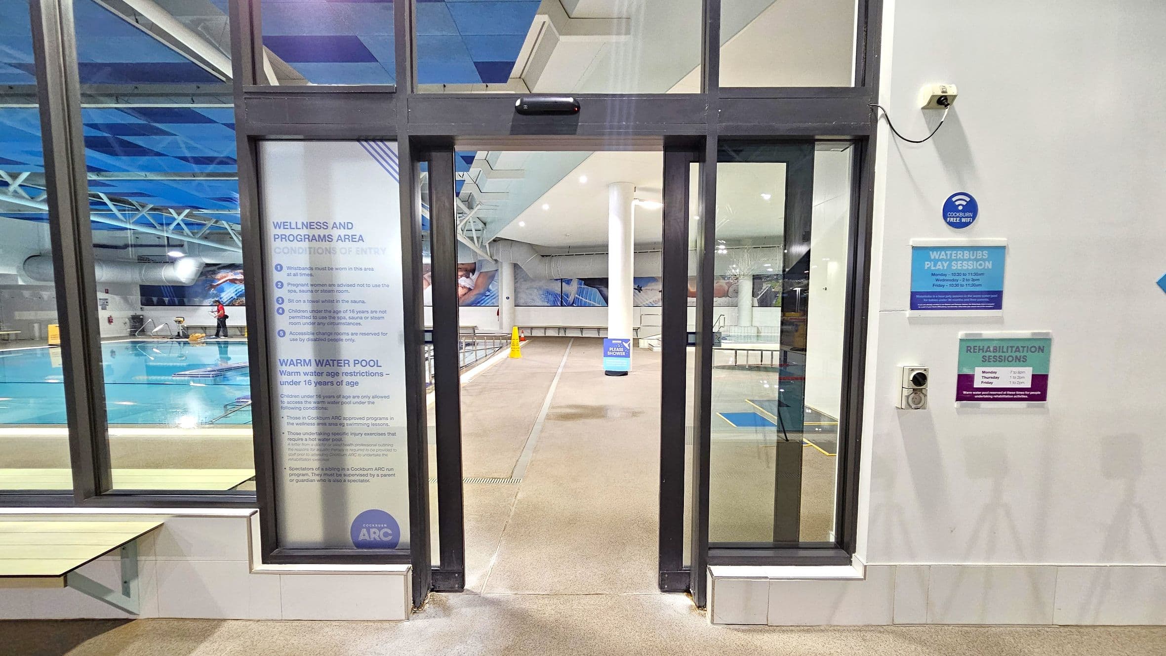 A glass entrance to a swimming pool area with signs on both sides. On the left, a large window shows a pool with a person cleaning it. A sign lists conditions of entry and age restrictions for the warm water pool. On the right, there are signs for "Waterbubs Play Session" and "Rehabilitation Sessions," along with a "Cockburn Free WiFi" sign. The floor is tiled, and the ceiling has a blue geometric pattern. A yellow caution cone is visible in the background.