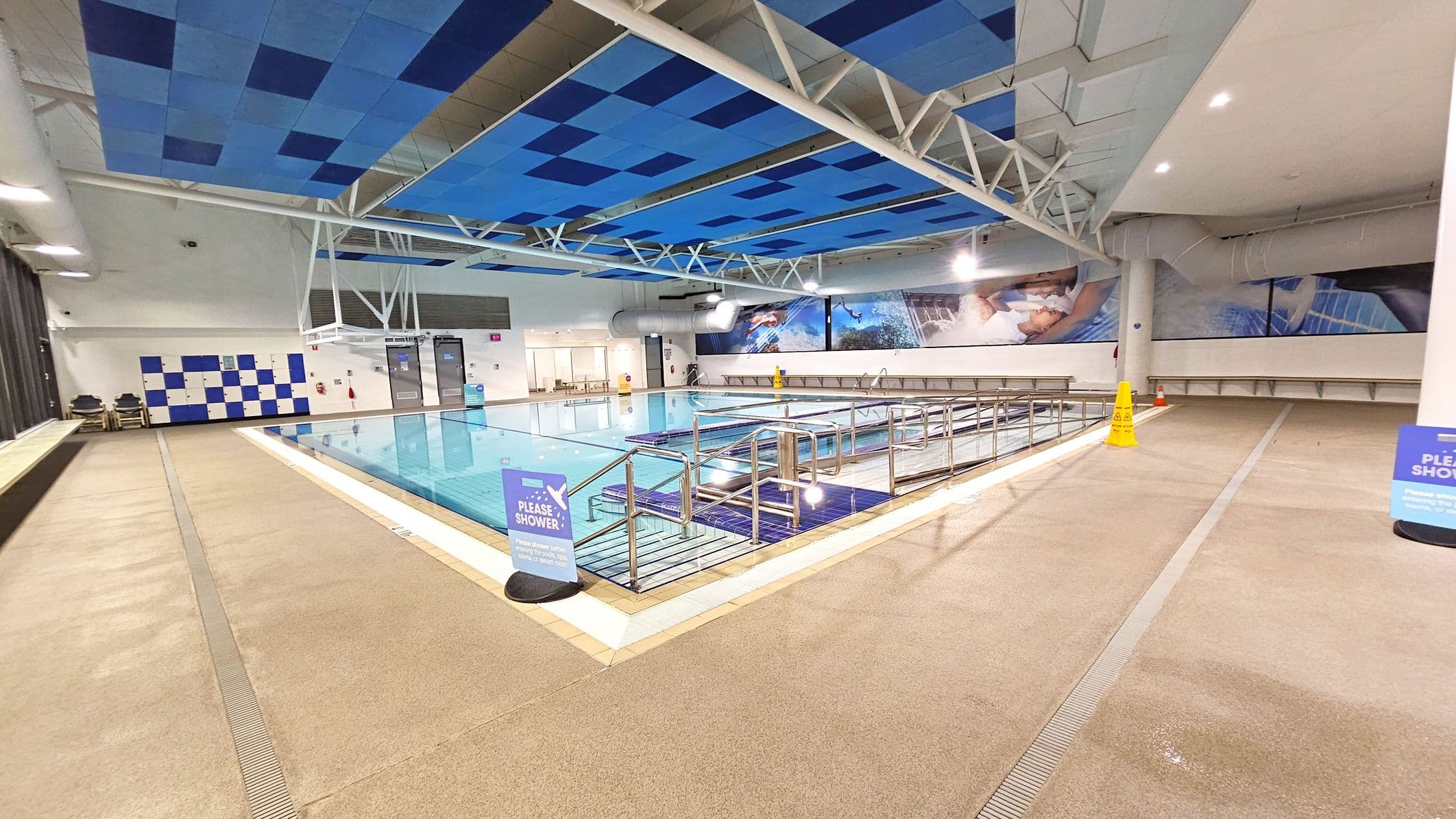 Indoor swimming pool area with a blue and white tiled ceiling. The pool is in the centre, surrounded by a beige tiled floor. On the left, there are lockers with a blue and white checkerboard pattern. On the right, a long bench runs along the wall, with a yellow caution sign on the floor. Large wall murals depict swimming scenes. A sign near the pool reads "PLEASE SHOWER." The ceiling features exposed white beams and blue panels.