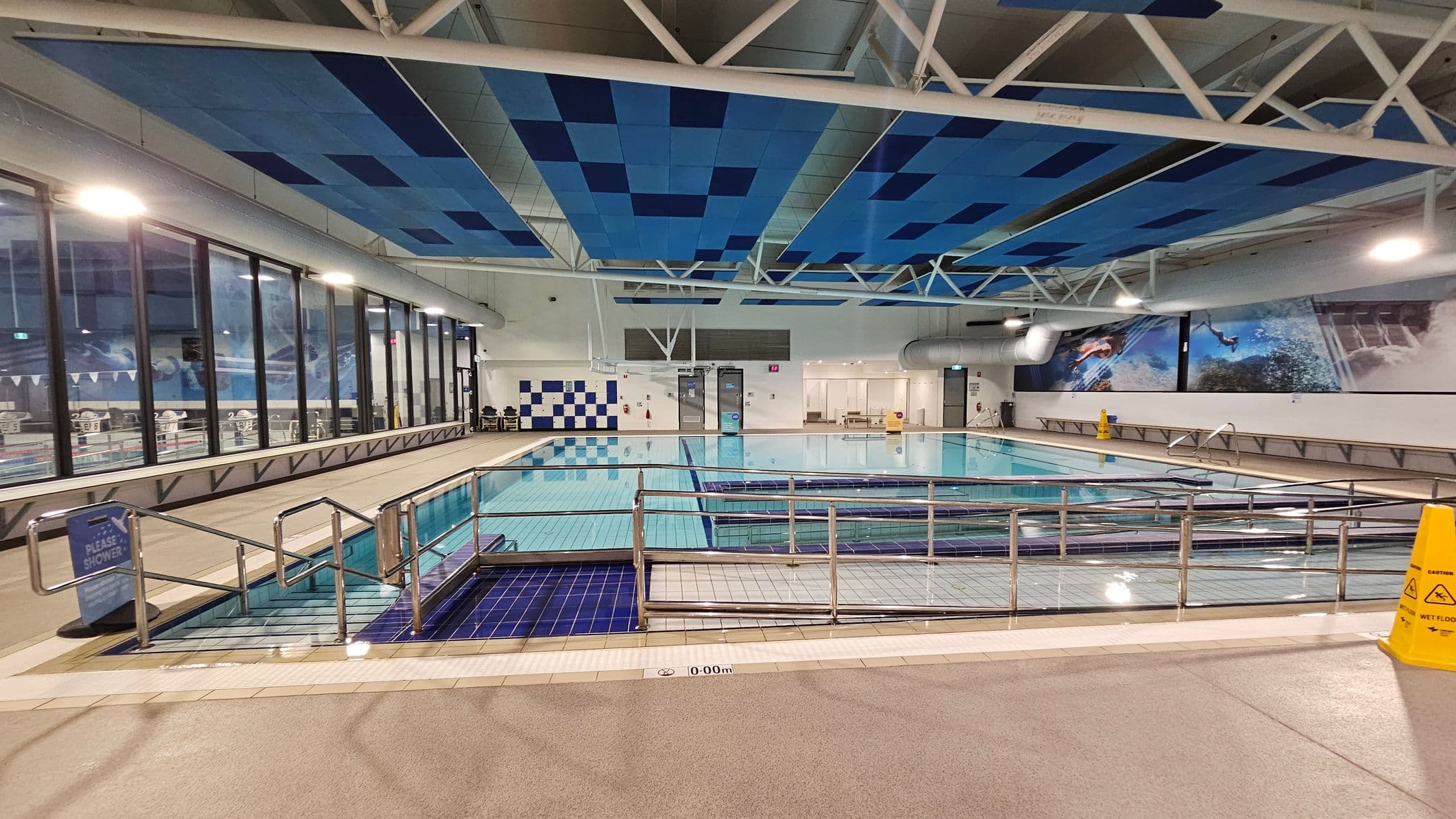 Indoor swimming pool with a ramp and handrails leading into the water. The left side features large windows with a view of another pool area. The right side has a wall with aquatic-themed murals. The ceiling is decorated with blue panels and white beams. The floor is tiled, with a wet floor sign visible. The pool area is well-lit with overhead lights.