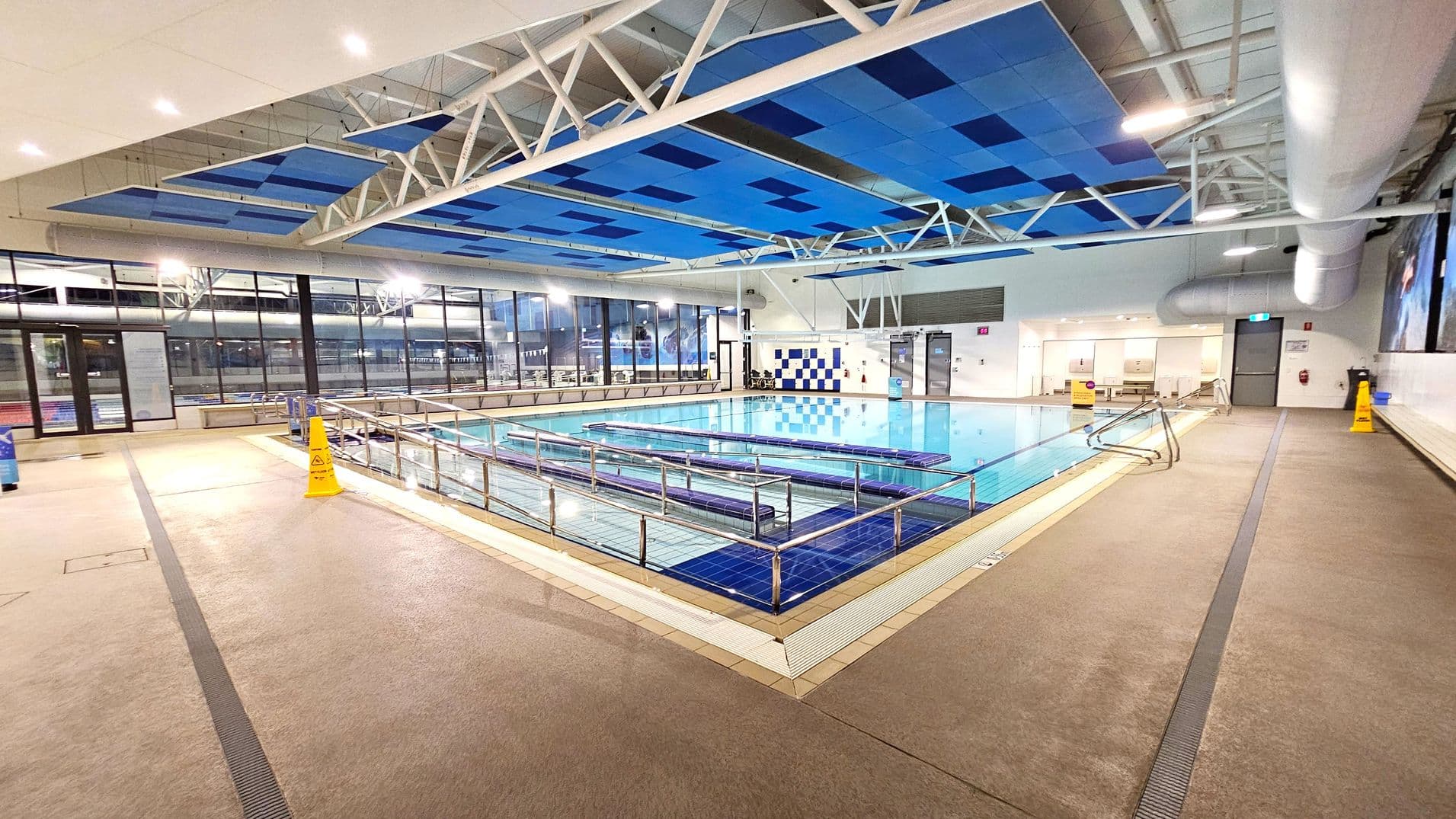 An indoor swimming pool with clear water, surrounded by a tiled deck. The ceiling features a geometric pattern of blue panels and white beams. Large windows line the left side, allowing natural light to enter. On the right, there are changing room doors and a large ventilation duct. Yellow caution signs are placed on the floor, which is a light brown colour. The pool has metal railings and a ramp for accessibility.
