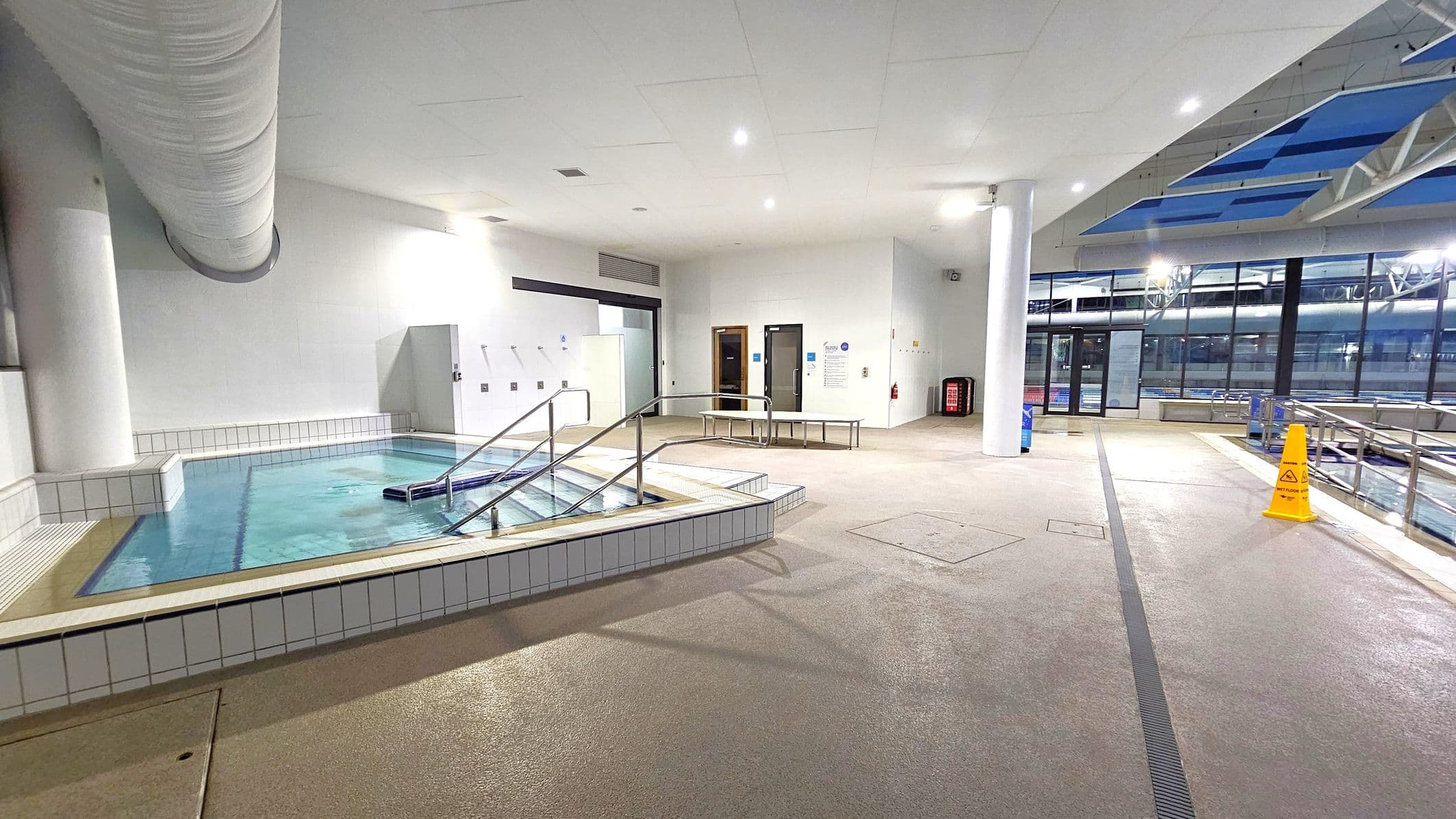 Indoor pool area with a small, square pool on the left, featuring metal handrails and steps leading into the water. The floor is light brown with a textured surface. A large white duct runs along the ceiling. On the right, there are large windows with a view of another pool outside. A yellow caution sign is placed near the windows. The ceiling is white with recessed lighting, and there are doors and benches along the back wall.