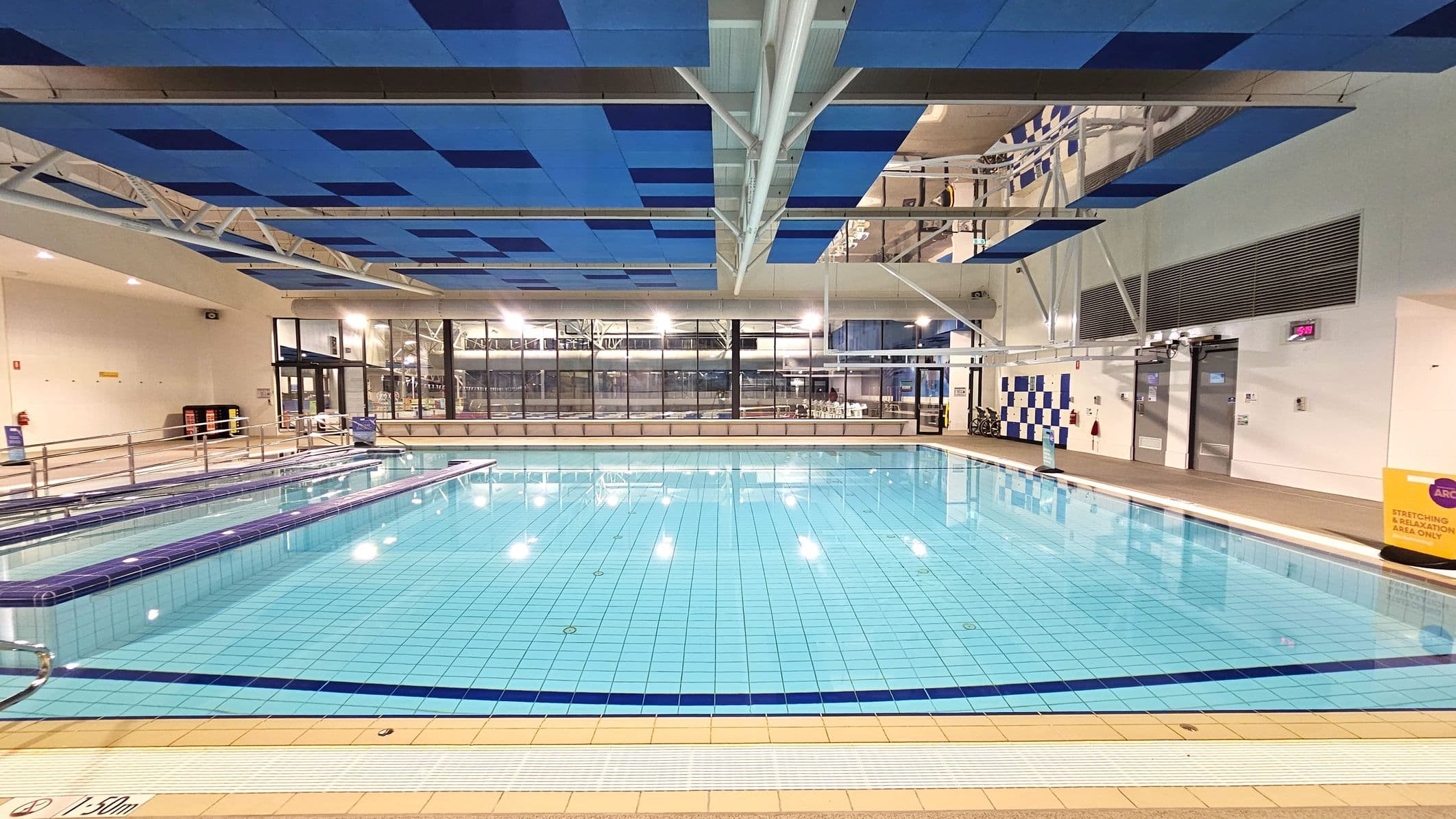 An indoor swimming pool with clear blue water and tiled flooring. The ceiling features a pattern of blue panels and exposed white beams. On the left, there are metal railings and a ramp leading into the pool. The background shows large glass windows with a view of the outside, and a wall with a blue and white checkerboard pattern. A digital clock and a yellow sign are visible on the right side. The pool area is well-lit with overhead lights.