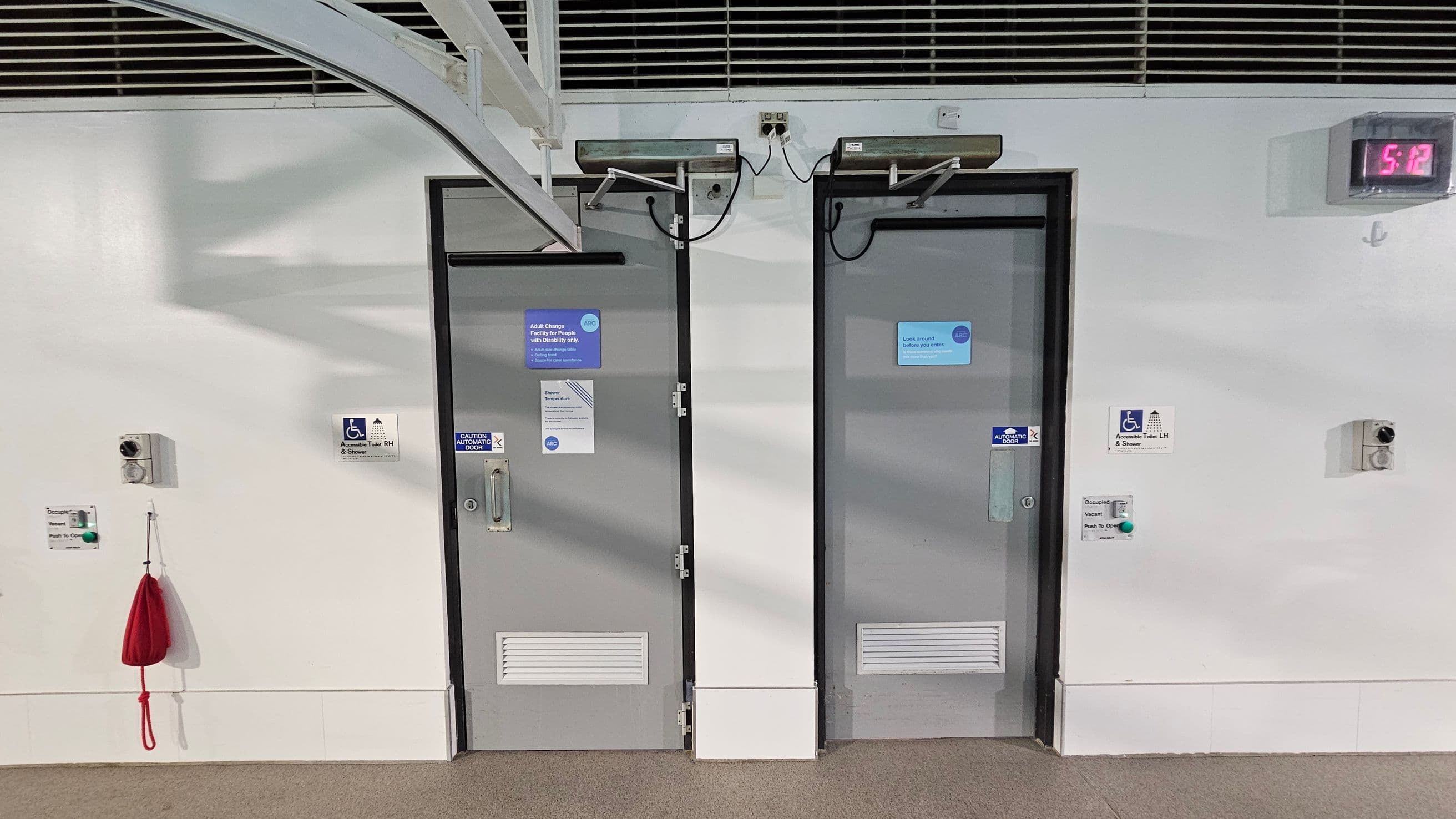 Two grey doors with signs, one on the left labelled for adult changing and the other on the right with a caution sign. Both have accessibility symbols. Above each door are automatic door mechanisms. A digital clock on the upper right shows 5:12. A red emergency pull cord hangs on the left wall, with a green button below it. The floor is carpeted, and the ceiling has metal beams and vents.