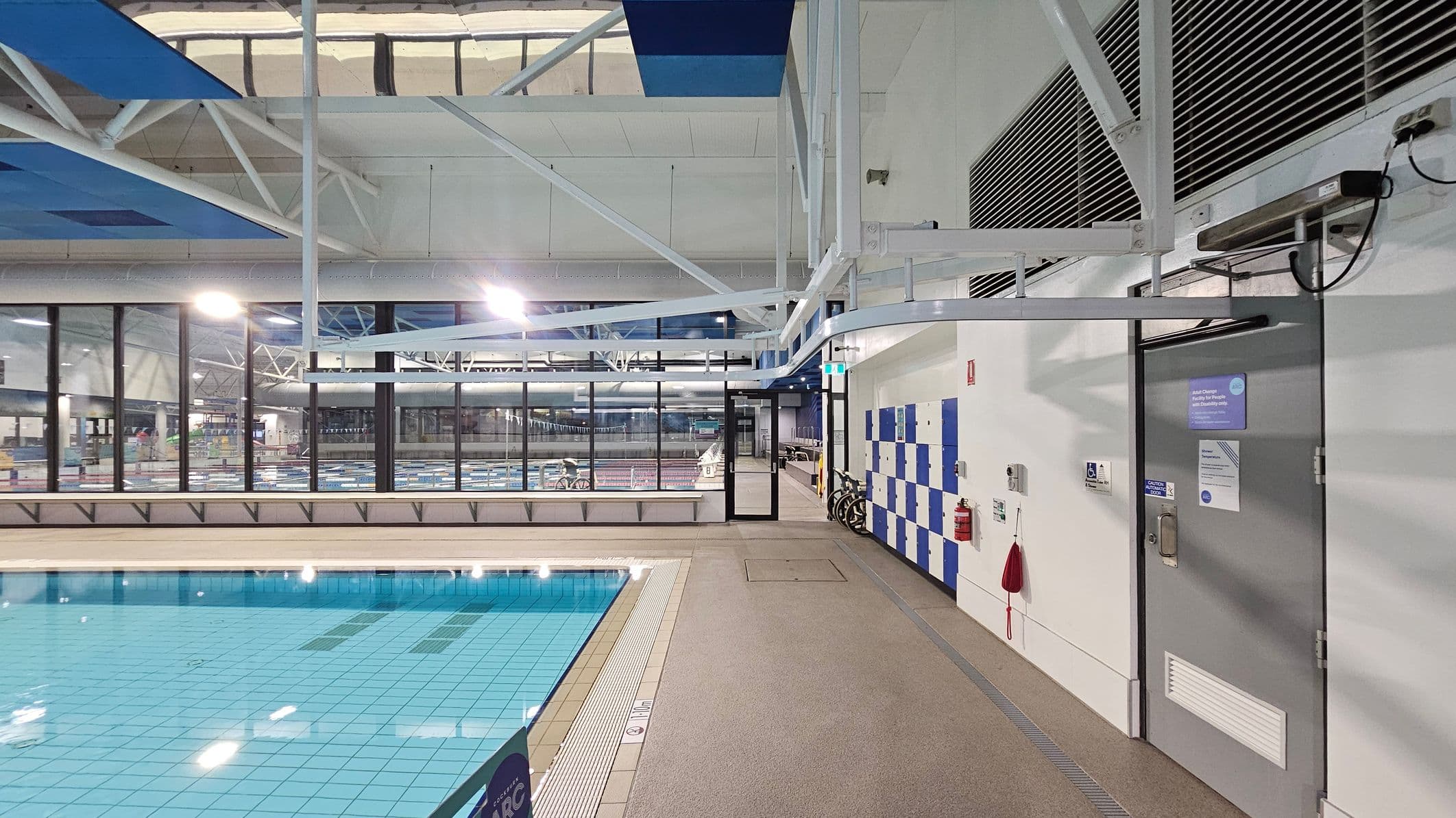 Indoor swimming pool area with a tiled pool on the left, featuring clear blue water. The right side has a wall with blue and white lockers, a grey door with signs, and a fire extinguisher. The floor is a non-slip surface, and the ceiling has exposed white beams and blue panels. Large windows in the background provide a view of another pool area with lane dividers. Bright overhead lights illuminate the space.