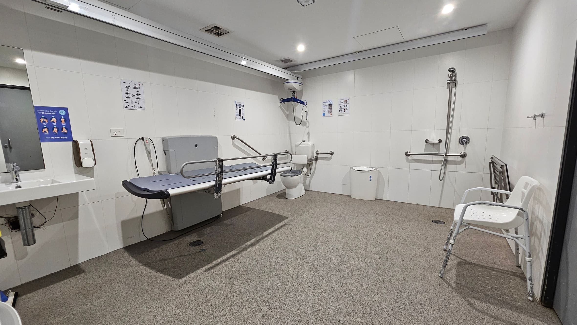 A spacious, accessible bathroom with white walls and a speckled green and brown floor. On the left, there's a sink with a mirror above it and a handwashing poster. A changing table with safety rails is mounted on the wall. In the centre, a toilet with support bars is visible, along with a ceiling-mounted hoist. On the right, there's a shower area with a handheld showerhead and a white plastic chair. The ceiling has recessed lighting.